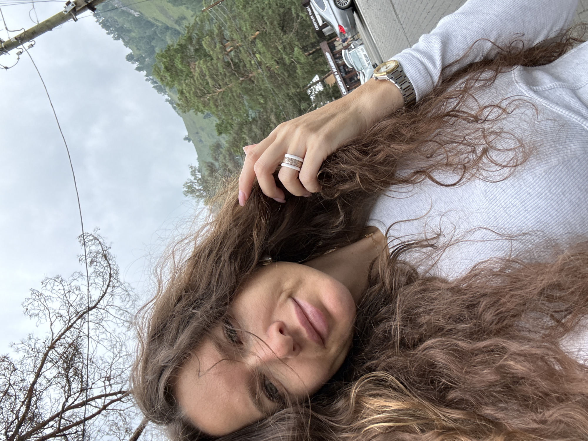 A woman with long, curly brown hair takes a selfie outdoors. She is wearing a white, long-sleeved shirt and a watch on her left wrist. Her left hand, adorned with two rings, is raised to her hair. In the background, a forested mountain slopes upwards under a cloudy sky. A street with parked cars and a building with Russian signage is visible to the right. The location is Manzherok, Russia.