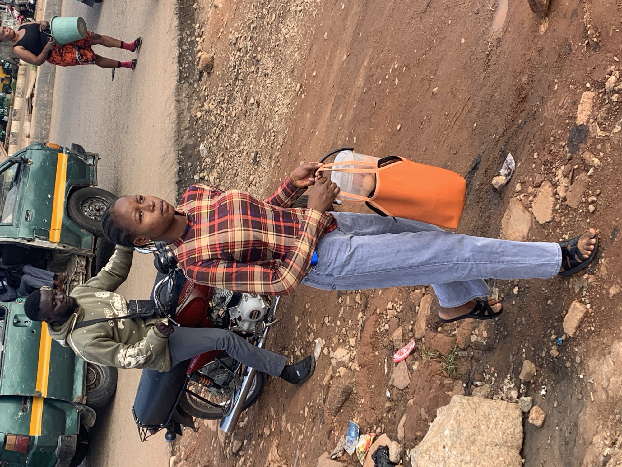 In Gura Top, Nigeria, a young woman walks towards the camera wearing a plaid shirt and light blue jeans. She carries a translucent bucket with a black handle in her right hand, partially covered by an orange carrier bag, containing what appear to be eggs. Her left hand is by her side. She is wearing black sandals. Behind her, a man on a red motorcycle, wearing a green hoodie and black pants, looks to his left. A green and yellow van is visible in the background. The ground is dirt and rocky, with scattered debris.