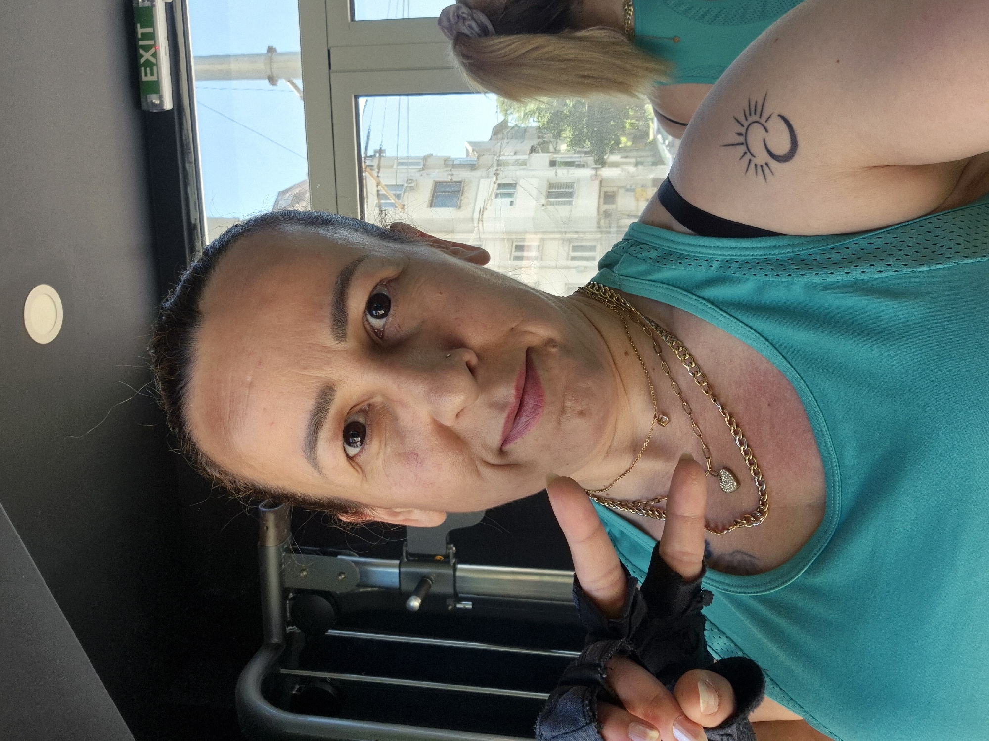 A woman is taking a selfie in what appears to be a gym. She is wearing a turquoise tank top and several gold necklaces. She has on fingerless gloves and is making a peace sign with her left hand. On her right arm, she has a tattoo of a crescent moon with a sun above it. In the background, there is a window looking out onto a city street with buildings. An "EXIT" sign is visible on the wall above the window.