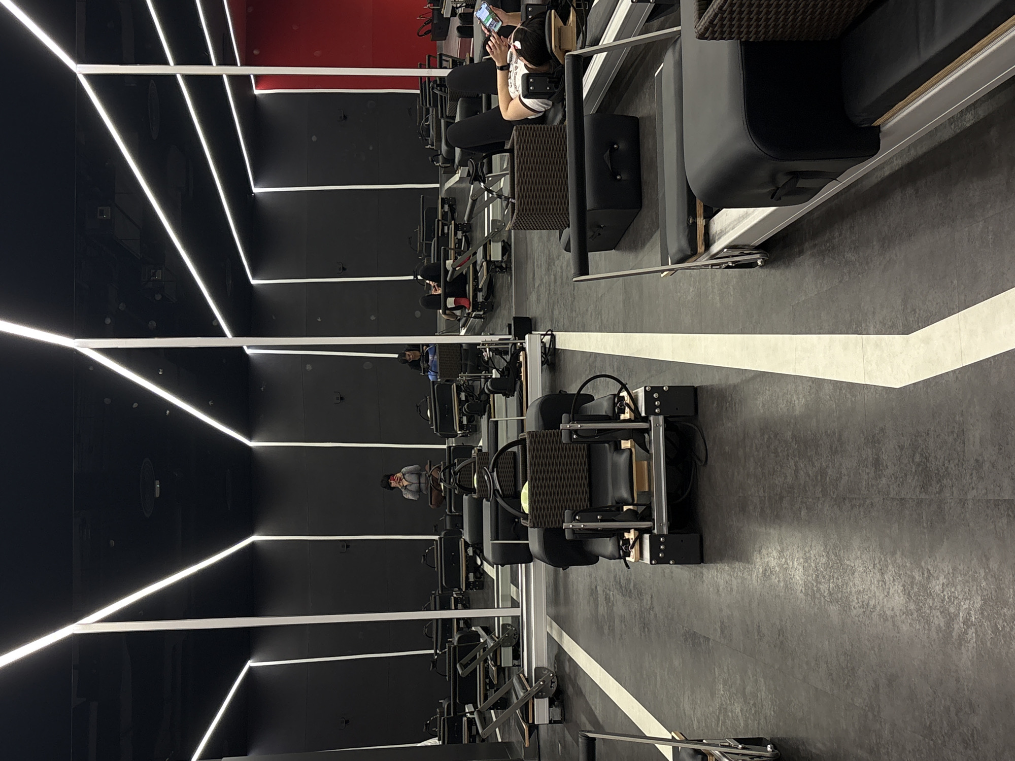 In a modern fitness studio with dark walls and ceiling, accented by linear white lighting, several pilates reformers are set up in rows on a dark grey floor marked with white lines. In the foreground, one reformer is equipped with a dark woven basket containing a yellow object. Further back, a person is lying on a reformer, looking at a mobile phone. Another person is seen in the mid-ground, taking a selfie with a phone. The studio appears to be empty of other patrons, with only a few individuals visible. The environment suggests a class or workout session is either about to begin, is ongoing, or has just concluded. There are reflections on the wall, indicating the presence of mirrors.
