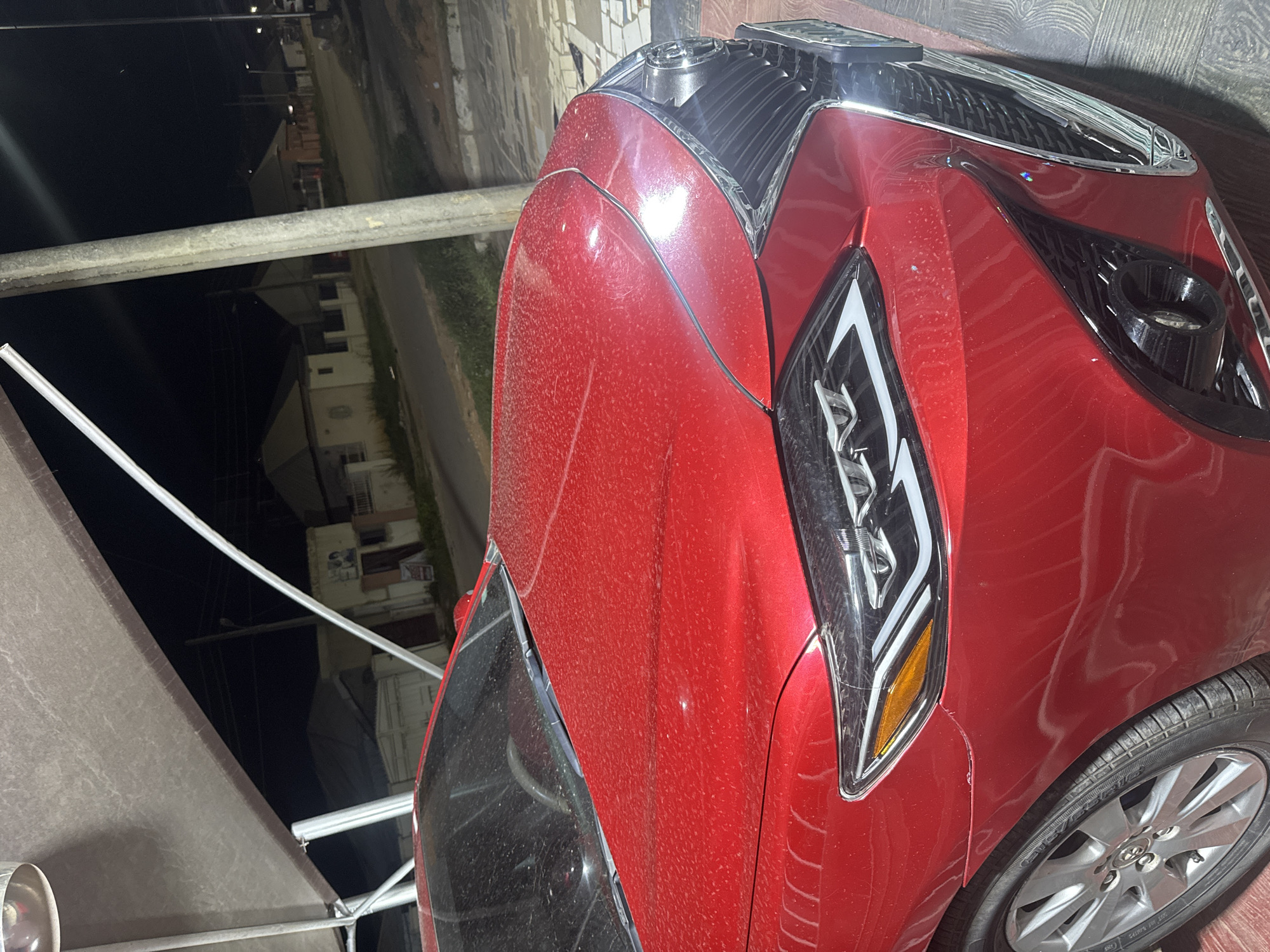 A red car is parked under a canopy at night in Abuja, Nigeria. The car's hood and windshield are speckled with dust or water droplets. In the background, houses with lights on are visible along a street. A concrete pole stands to the right of the car.