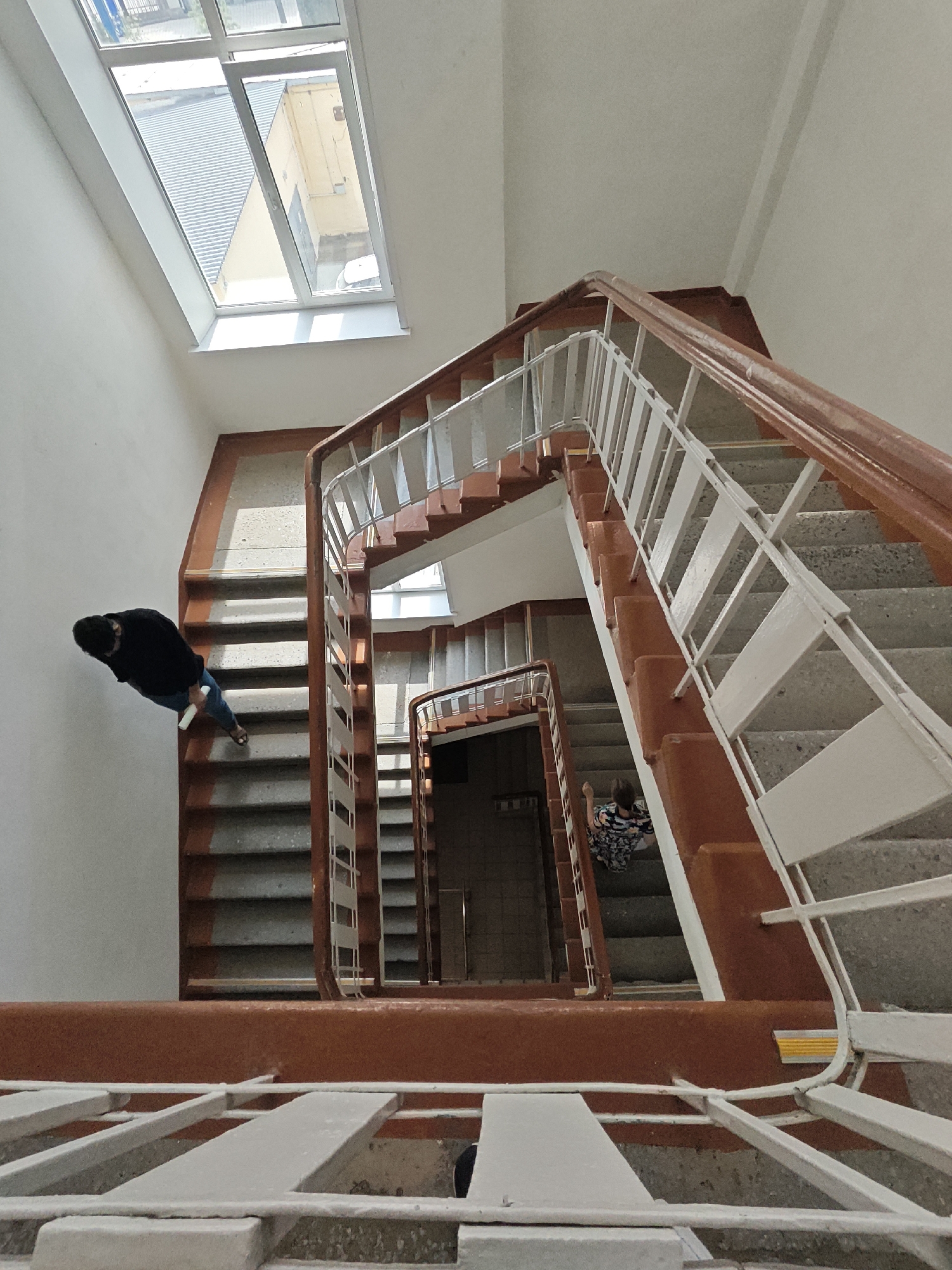 An eye-level, downward angled photograph depicts a multi-story staircase, constructed of concrete steps with terra cotta colored risers, within a building in Yekaterinburg, Russia. The staircase winds downward in a square spiral. The stair railing consists of a wooden handrail painted terra cotta and white painted metal bars. Two people are present. A woman wearing a dark top and blue jeans with a white object in her right hand walks down the steps on the left side of the frame. A second person, also a woman, wearing a patterned dress and light colored shoes, is descending the stairs on the right side of the frame. A window is visible on the top left. The walls are painted white. A doorway is visible in the background. The stairs lead to different levels, suggesting a multi-story building.
