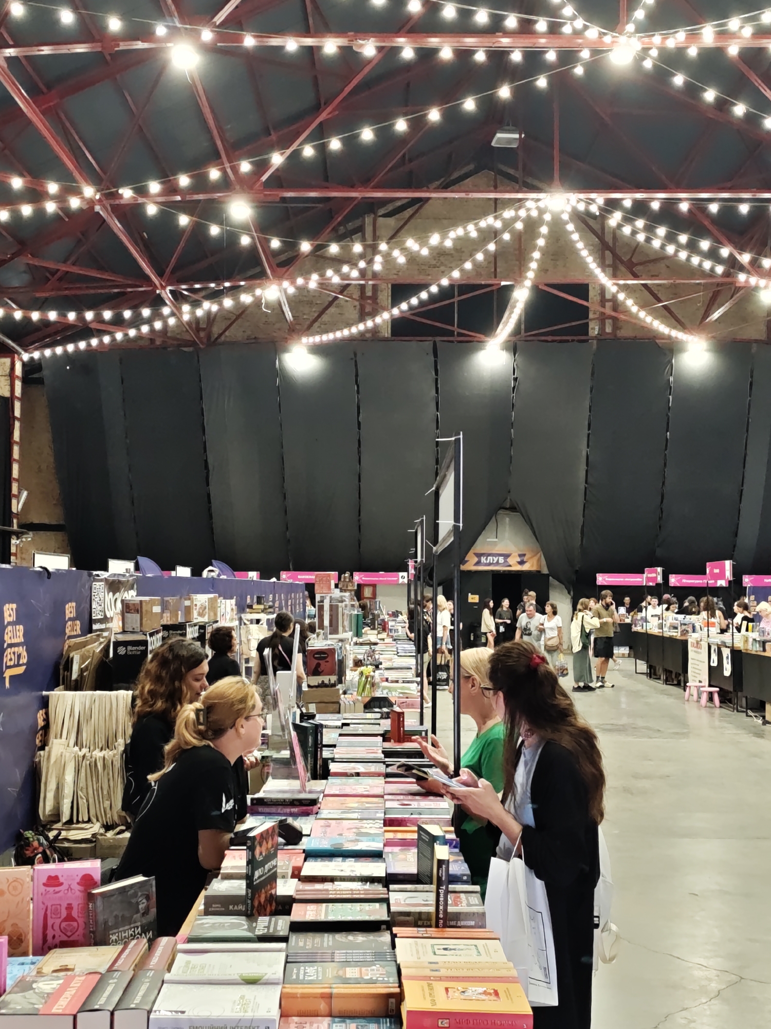 The image shows an indoor book fair, likely located in Lviv, Ukraine, given the Cyrillic text visible on some book covers and signage. The scene depicts a large, open space with a high ceiling adorned with strings of lights. Several people are browsing tables laden with books. In the foreground, two women are looking at books displayed on a table. One woman, wearing glasses and a black t-shirt, is leaning over the table, while the other, in a black dress, is holding a phone and a shopping bag. Behind them, more people are visible, along with numerous bookshelves and displays of books. The overall atmosphere suggests a lively event focused on literature and reading.