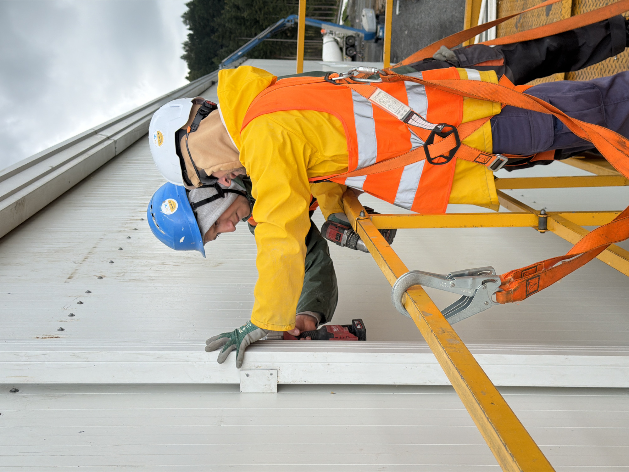 Two individuals, wearing hard hats and safety vests, are engaged in construction work on a white, corrugated building facade. The person on the left, wearing a blue hard hat and a green jacket, is holding a black and red power drill, appearing to be screwing into the wall. The person on the right, with a white hard hat and a yellow hooded jacket, is wearing an orange safety vest and is also positioned near the wall. Both individuals are on a yellow safety platform, secured by an orange harness with a metal clip attached to the railing. The background shows a cloudy sky, trees, and construction equipment, including a blue boom lift and a white concrete truck. The image is a medium shot, capturing the upper bodies and immediate surroundings of the two workers.