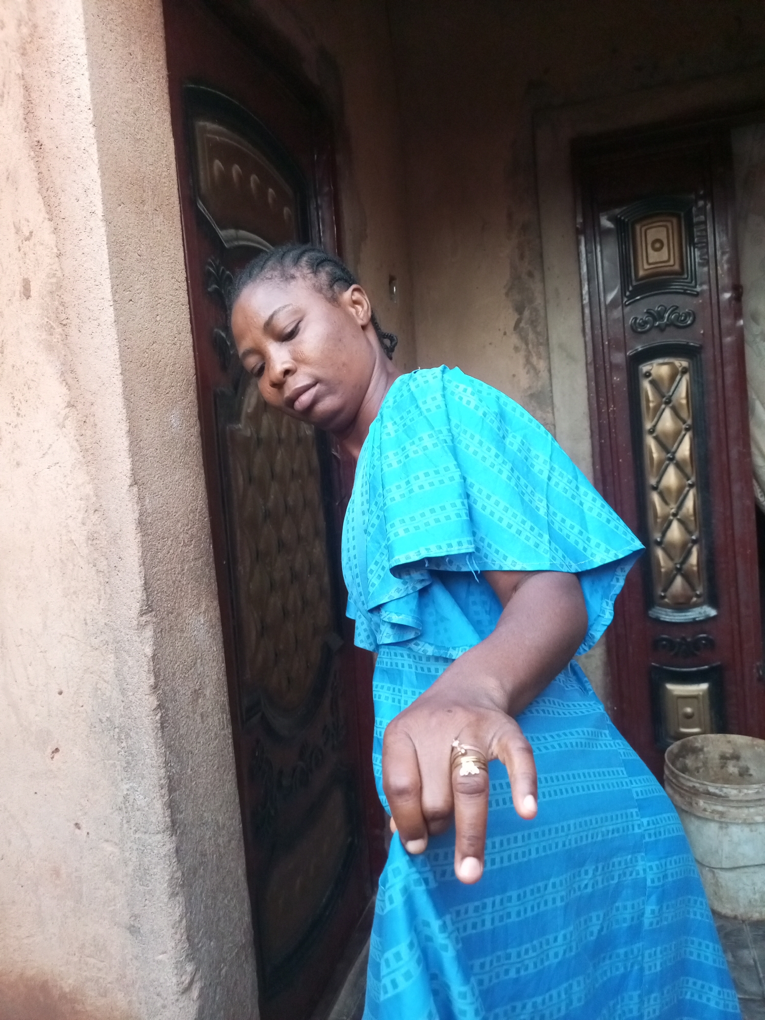A young Black woman with braided hair leans against a textured, tan-colored wall next to a dark brown, ornate door. She is wearing a bright blue dress with a ruffled, layered design and a geometric pattern. Her head is tilted down, eyes closed, and her lips are slightly parted. Her right hand is extended towards the viewer, with two fingers pointing upwards in a "peace" sign. A gold ring with a floral design is visible on her ring finger. To the right of the door, a light gray plastic bucket sits on the ground. The background consists of the wall and the door, suggesting an outdoor or entryway setting in a residential area.