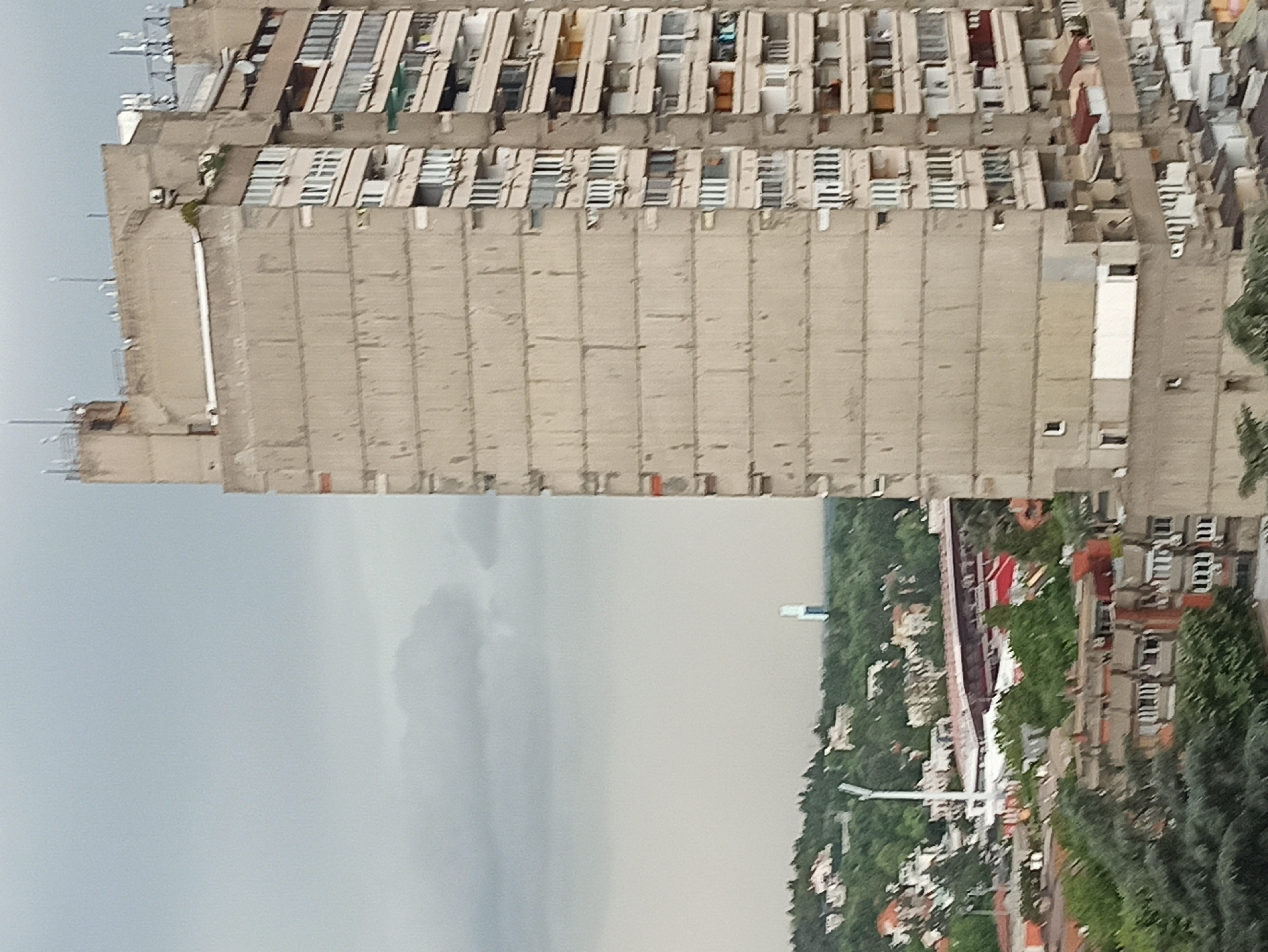 The image shows a tall, rectangular concrete building in Vozdovac Urban Municipality, Serbia. The building has a repetitive pattern of windows and balconies on its long side. In the background, a city landscape with buildings, trees, and roads can be seen. The sky is overcast with gray clouds. The image orientation is rotated.