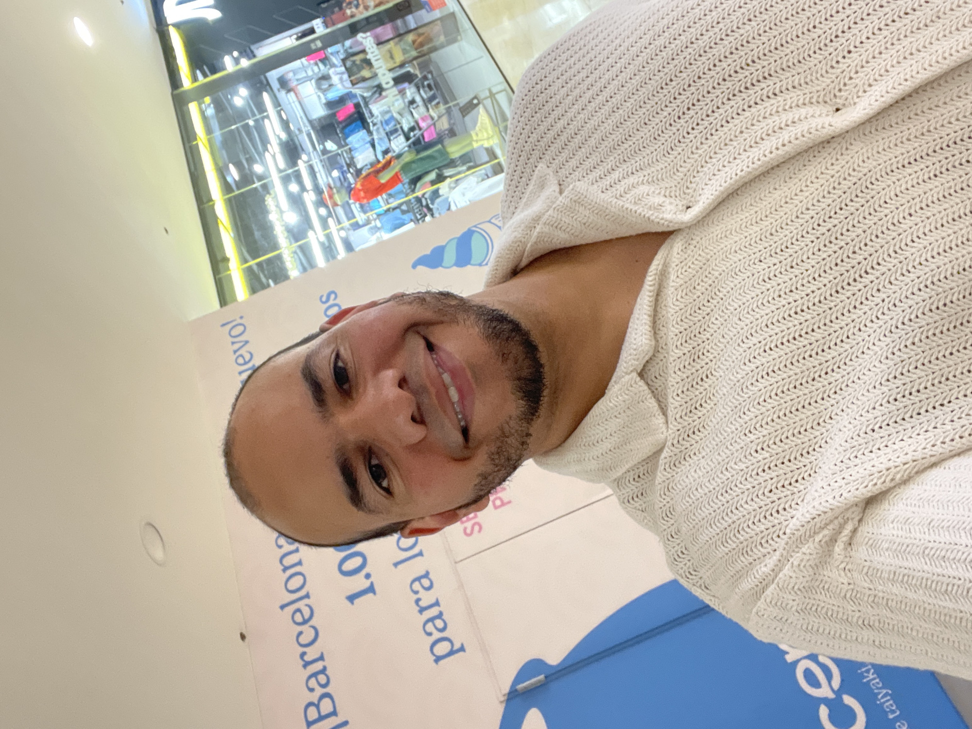 A man with a beard and a light-colored, knitted shirt smiles at the camera in a selfie taken indoors. Behind him, a large banner with blue text reads "Barcelona 1.0" and "para lo" in Spanish. To the right of the man, through a glass partition, is a store with mannequins and clothing displays, illuminated by bright lights. The ceiling above has circular lights.