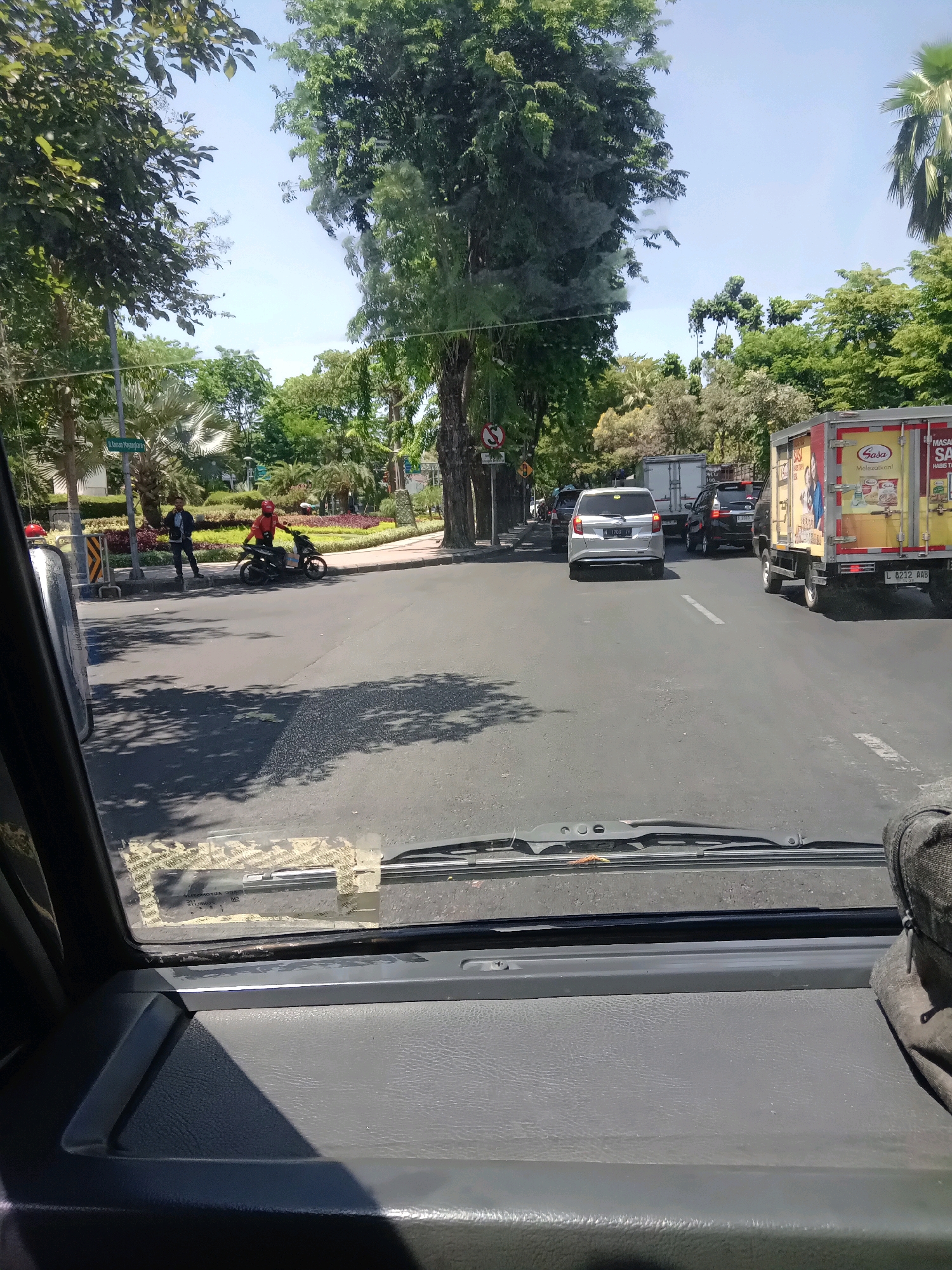 The image captures a street scene in Surabaya, Indonesia, viewed from the dashboard of a vehicle. The foreground shows a gray dashboard and a windshield with reflections and what appears to be a sticker taped to the interior. Outside the windshield, traffic is visible on a sunny day. Several cars, including a silver hatchback and a black SUV, are in motion or stopped in traffic. A large yellow truck with "Sasa" branding is prominent on the right side of the road, displaying text in Indonesian. On the left side of the road, a motorcycle with two individuals is stopped. One person is standing next to the motorcycle, and another is seated on it, wearing a red helmet. Trees line the street, and a manicured median with plants and a sign reading "Jl. Darmo Mapanjaja" is visible. The sky is clear and blue.