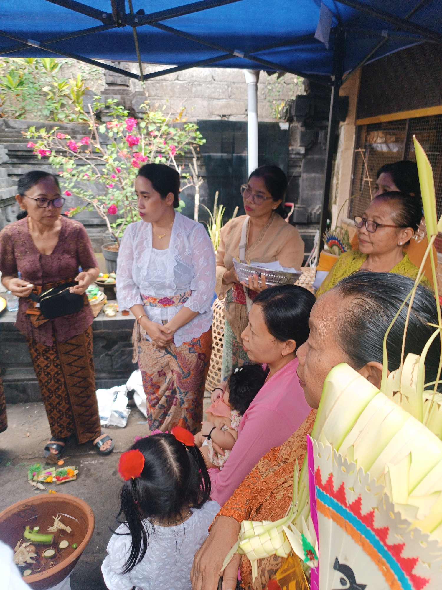 In an outdoor, covered area in Tabanan, Indonesia, a group of at least seven individuals, primarily women and a child, are gathered. The setting appears to be a courtyard with stone walls and a blue tarp overhead. Some individuals are dressed in traditional Balinese attire, including sarongs and kebaya-style tops. One woman, on the left, wears a brown patterned sarong and a dark brown top, with glasses and a black bag slung across her body. Next to her, a woman in a white lacy kebayas and a patterned sarong stands with her hands clasped. Another woman, to the right of the first, wears a light beige kebayas and holds a silver tray. Further to the right, a woman in a yellow kebayas looks on, and in the foreground, a woman in a bright pink top and an orange lace garment is visible, holding a baby. A young child with dark hair and two red hair accessories is in the lower foreground, facing away from the camera, wearing a white top. On the ground, there are offerings made of leaves and colorful decorations, as well as a brown clay bowl containing various items, including a cucumber slice. The overall atmosphere suggests a cultural or religious gathering.