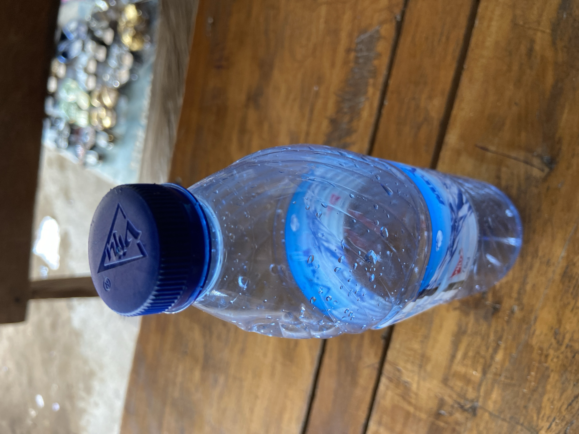 An eye-level shot shows a clear plastic water bottle with a blue cap on a wooden table. The bottle is partially filled with water, and condensation has formed on its exterior. The label on the bottle is partially visible, featuring blue and white designs. The wooden table has a natural grain and appears worn. In the background, the scene is blurred, suggesting a shallow depth of field, but hints of other objects and possibly a floor are discernible. The location is described as Mararaba, Nigeria, though no specific landmarks or contextual details within the image directly confirm this.