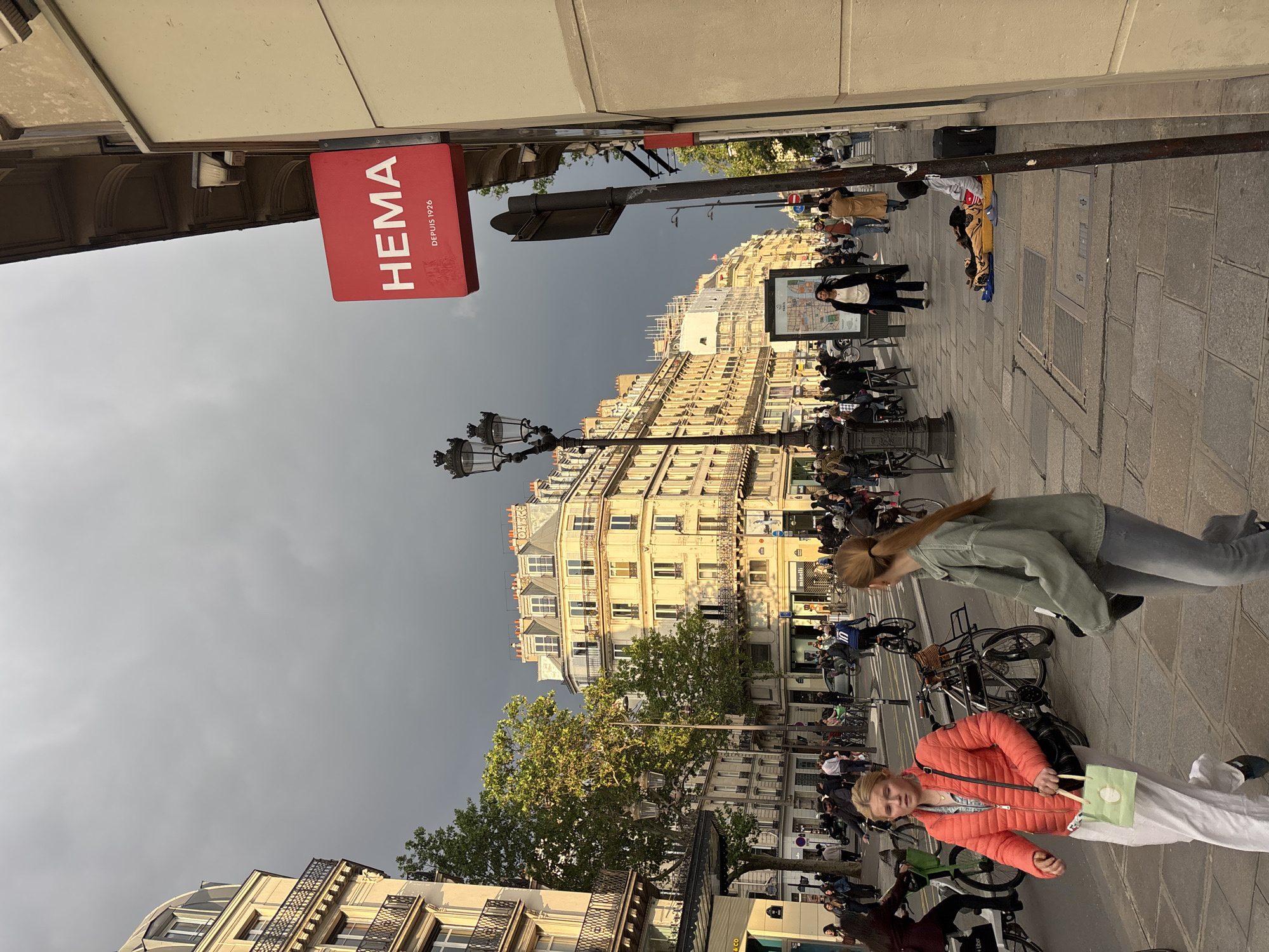 The image shows a street scene in Paris, France. A red "HEMA DEPUIS 1926" sign is visible on the left side of the frame. The street is crowded with people, bicycles, and street furniture. The architecture is typical of Parisian buildings with multiple stories and ornate facades. The sky is overcast. People are walking and interacting on the sidewalk. Some people are on bicycles. One person is lying on the sidewalk.