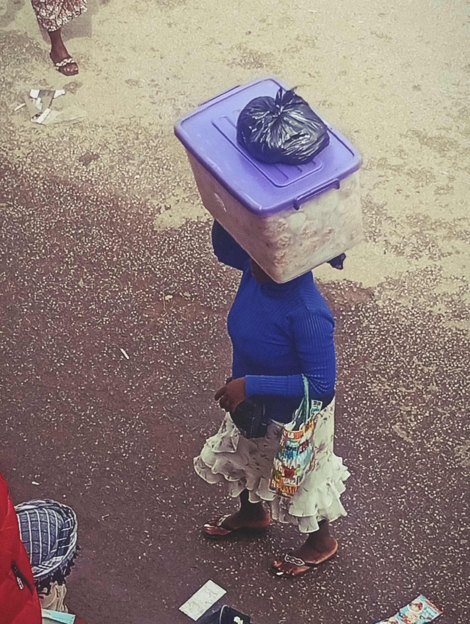 In Bukuru, Nigeria, a woman walks across a textured, outdoor ground surface, carrying a large, translucent plastic container filled with items on her head. A black plastic bag is tied and placed on top of the container. The woman is wearing a long-sleeved blue top and a layered, ruffled white skirt. She has a colorful fabric bag slung over her shoulder and is wearing brown flip-flops. The ground is dark and appears to be asphalt or concrete with small, scattered white debris. In the background, to the left, a person's legs are visible, wearing a patterned skirt and sandals. To the lower left, another person is partially visible wearing a red top and a striped headscarf.