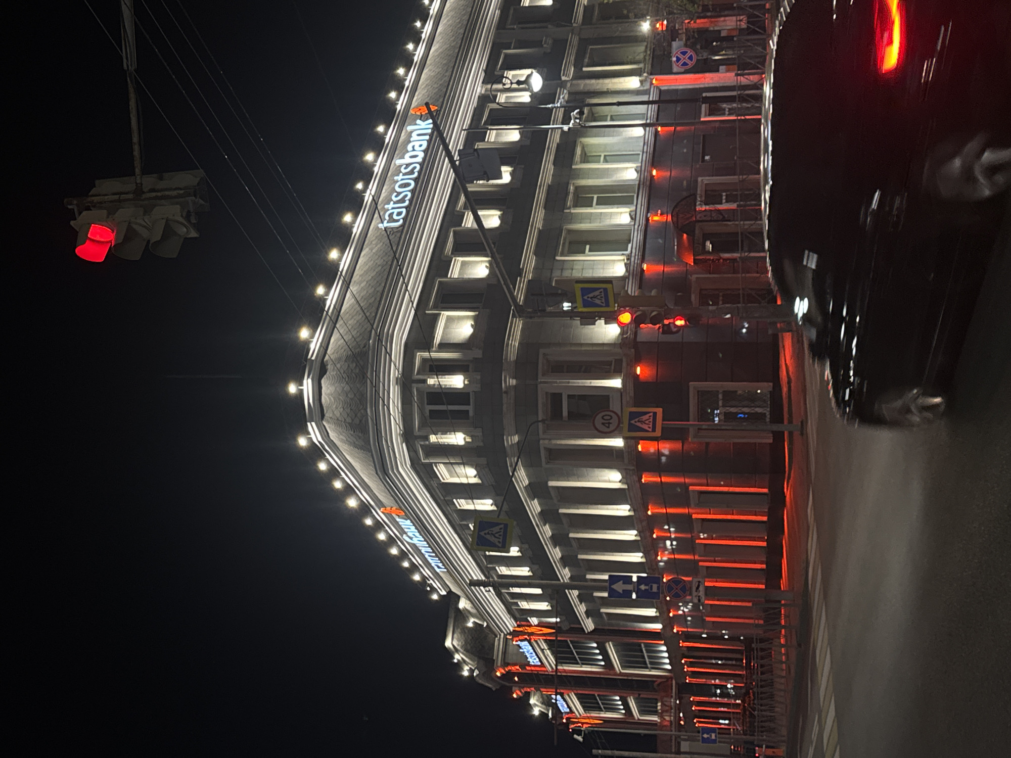 The image captures a night scene in Kazan, Russia, focusing on a building with illuminated signage for "tatsotsbank". The building features decorative red lighting along its facade and is topped with illuminated string lights. A traffic light at a crossroads is clearly visible, displaying a red signal. A black car is in motion in the foreground, partially blurring the lower portion of the image, with its taillights illuminated. Several street signs, including pedestrian crossing and speed limit signs, are present. The overall atmosphere is urban and nocturnal, with the lights of the building and traffic signal dominating the scene against the dark sky.