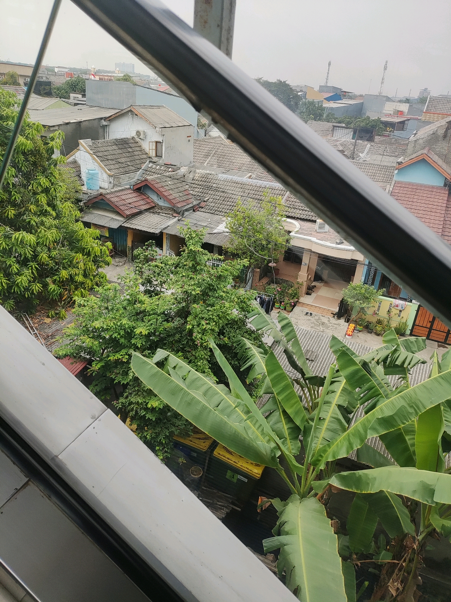This is an aerial shot of a residential area in Telagamurni, Indonesia, taken from a higher vantage point. The view is partially obstructed by a metal railing in the foreground. The scene displays a dense cluster of houses with tiled roofs, predominantly in shades of gray and terracotta. Lush green trees, including a prominent banana plant with large leaves in the lower right quadrant, are interspersed among the buildings. Clothes are seen hanging out to dry in one courtyard. The overall atmosphere suggests a typical urban neighborhood with a mix of residential structures and vegetation. The sky is overcast, indicating a possibly humid or cloudy day.