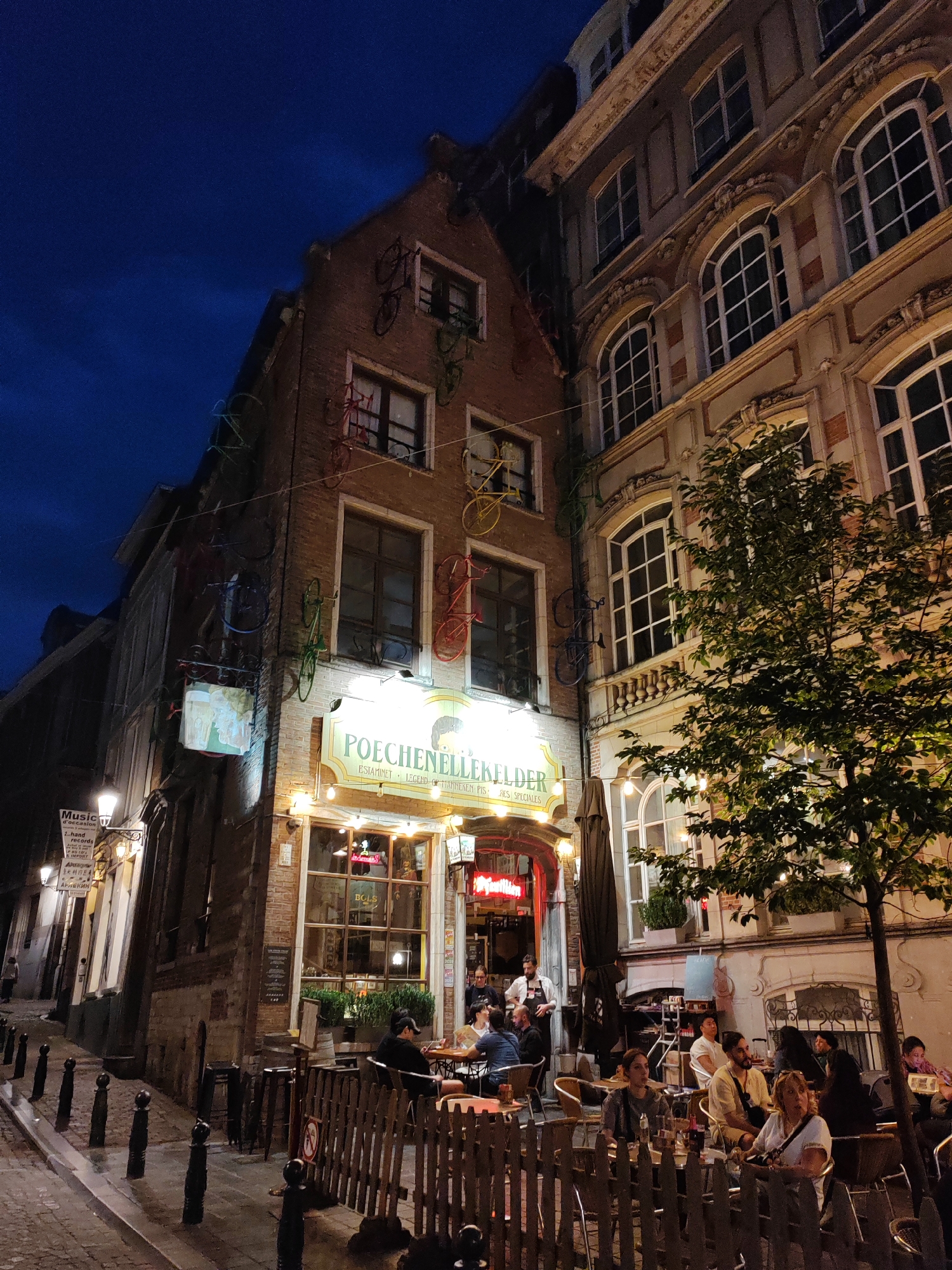 This image depicts a street scene in Brussels, Belgium, at twilight. The focus is on a building adorned with several vintage bicycles mounted on its brick facade. The ground floor of this building features a brightly lit establishment with a sign that reads "POECHENELLEKELDER ESTAMINET LEGEND OF HANDGEKER PIES." People are seated at tables outside this establishment, some interacting with each other, while others appear to be waiting to be served. The street is paved with cobblestones and lined with bollards. Adjacent to the establishment is another building with classical architectural elements and large arched windows. A tree with green foliage stands in front of this building, its branches illuminated by string lights. The sky is a deep blue, indicating the transition from day to night.
