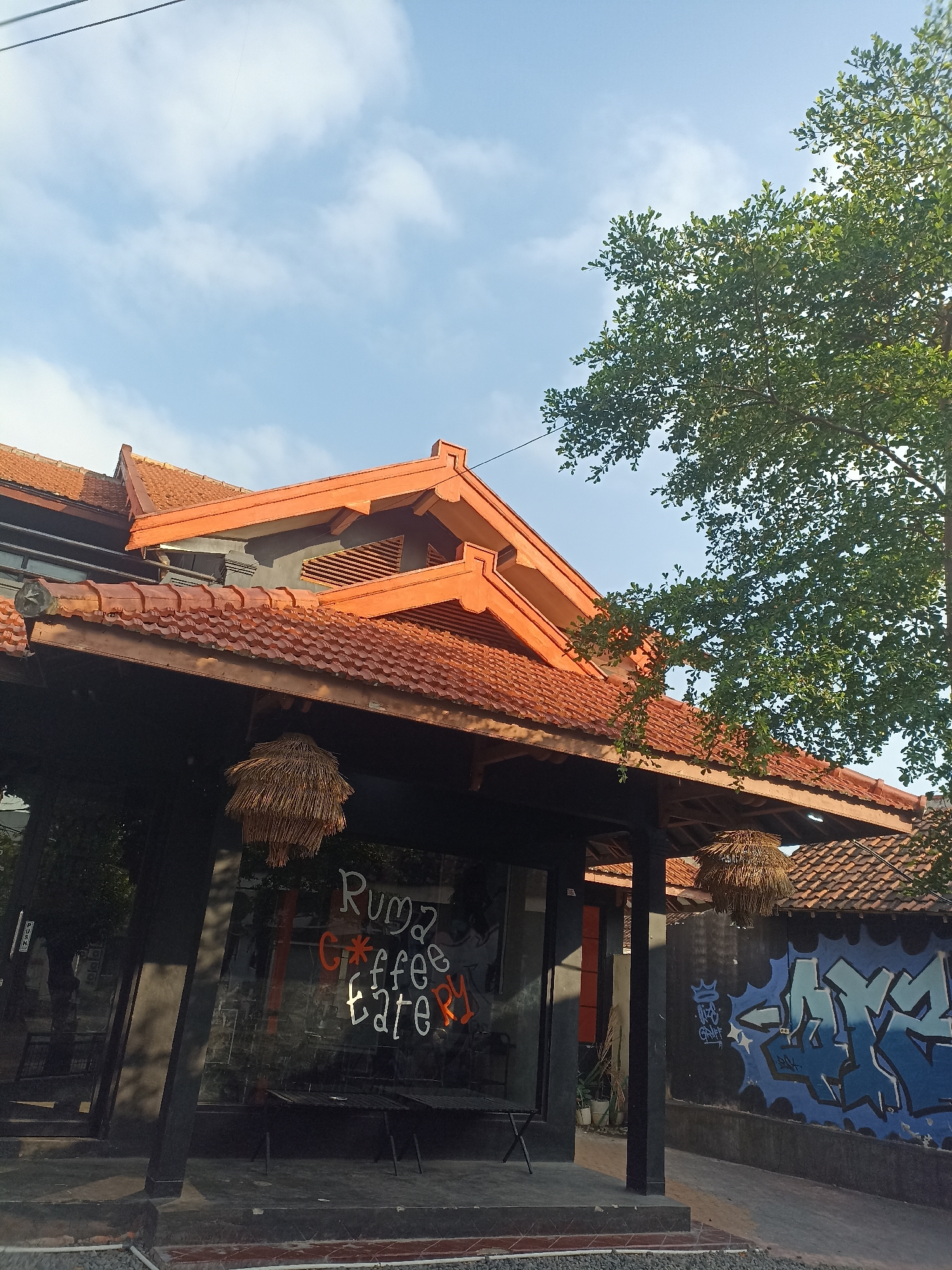 The image depicts a building with a distinctive orange tiled roof, possibly a coffee shop or eatery, in Banyuwangi, Indonesia. The building features dark-colored walls and large glass windows. The text "Ruma G*ffee tatery" is visible on one of the windows, rendered in white and orange. Two straw-like decorative elements hang from the eaves. To the right of the building, a concrete wall is adorned with blue graffiti. A lush green tree with dense foliage is visible in the upper right portion of the frame, with its branches extending towards the sky. The sky is clear blue with a few scattered white clouds. The overall impression is that of a bright, possibly sunny day. The scene is urban or semi-urban, given the presence of a building, wall, and what appears to be a paved walkway or driveway. No people are clearly visible in the image.