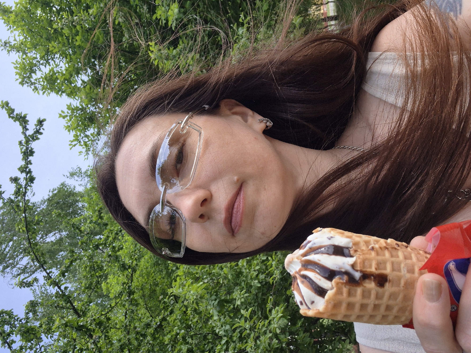 A woman with long brown hair and clear-framed glasses holds an ice cream cone in her hand. The ice cream is striped with chocolate sauce. The background shows lush green trees. The location is Unknown, Unknown.