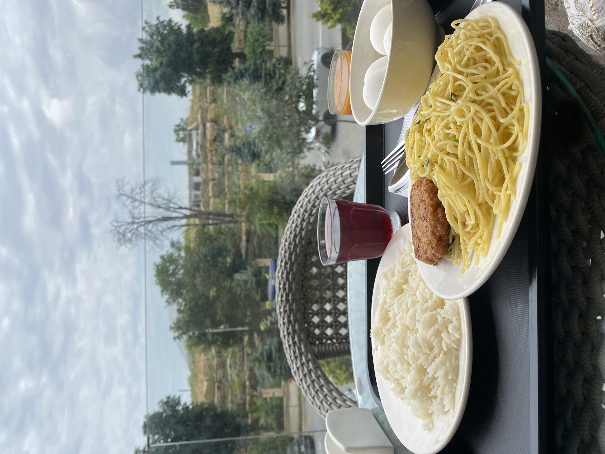 A close-up, eye-level shot shows a tray of food set against a blurred outdoor scene. The tray holds a plate of white rice, a plate of spaghetti with a small patty, a bowl of hard-boiled eggs, and two glasses of juice, one red and one orange. In the background, a wicker chair is visible, along with a window that overlooks a hillside with trees and a road. The overall setting suggests a restaurant or cafe with a view. The outdoor scene is likely Derbent, Russia, given the distinctive hillside landscape and architecture visible in the background.