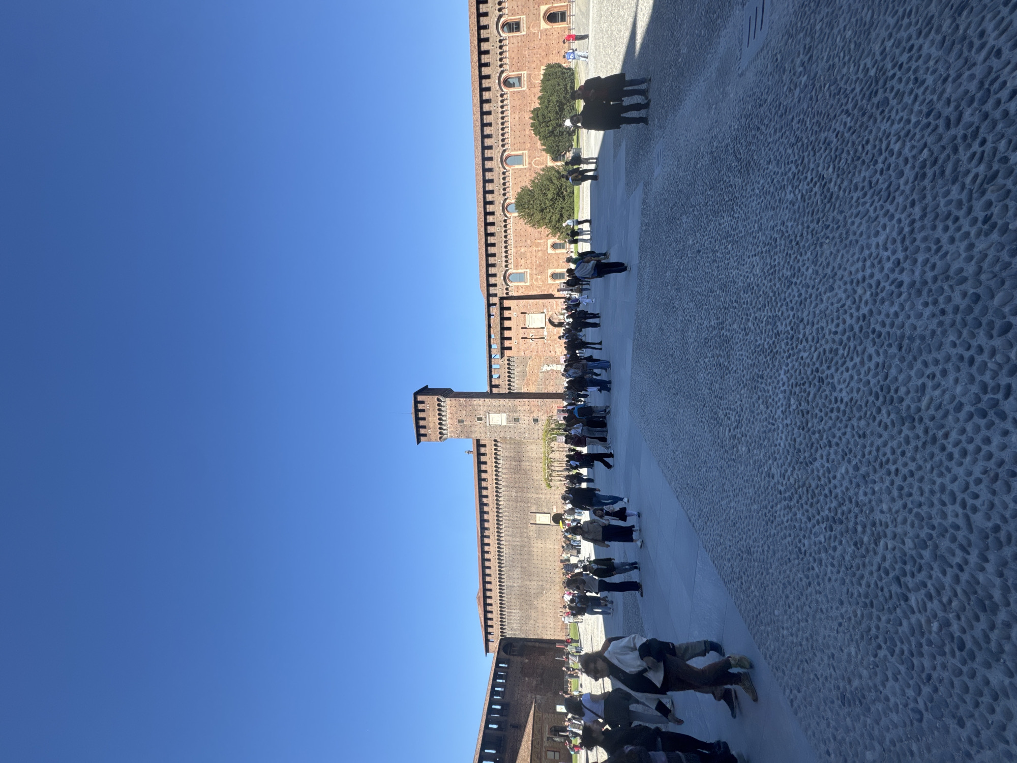 The image depicts Sforza Castle in Milan, Italy, on a sunny day. The castle's brick walls and turrets are visible, with numerous people walking and standing in the courtyard in front of the castle. The courtyard is paved with small stones. The sky is clear and blue.