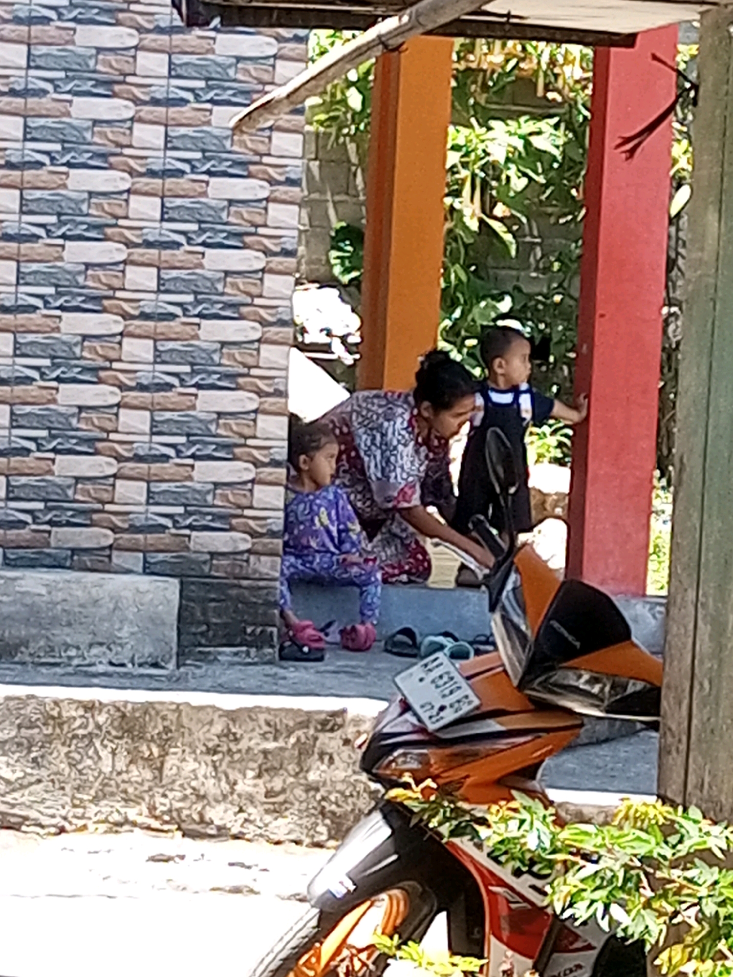 In Andongsili, Indonesia, a woman is tending to a young child who is looking away from the camera. Another young child sits on a step to the left. They are on a porch with a tiled wall and an orange and red pillar. A motorcycle is parked in the foreground.