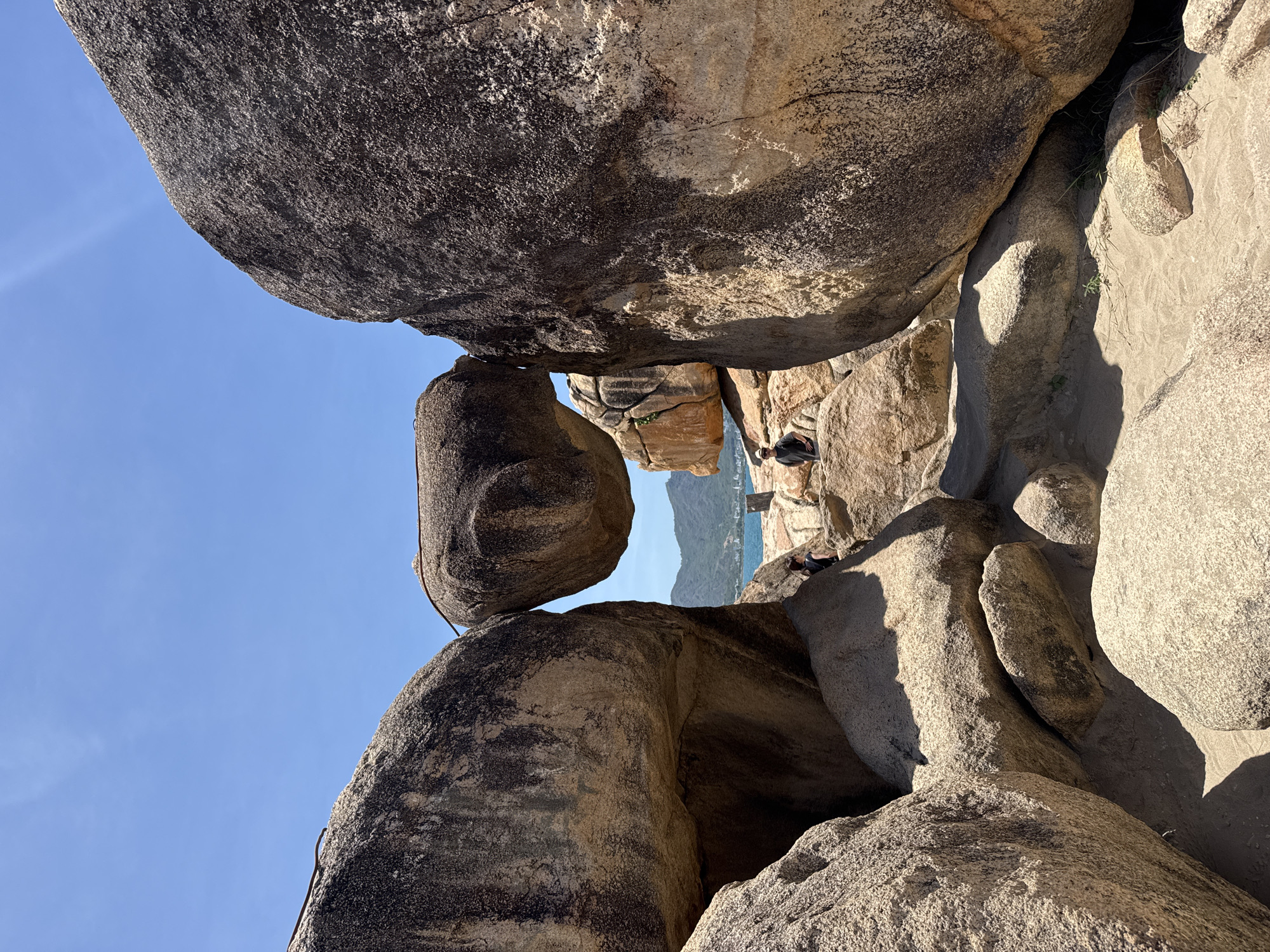 This photograph shows a cluster of large, weathered rocks in the city of Bac Nha Trang Ward, Vietnam. The rocks are stacked and arranged in a haphazard manner, creating narrow openings and shadowed areas. Two people are visible within the rock formation, their presence dwarfed by the surrounding stone. In the background, a glimpse of the sea and a distant mountain range is visible through the gaps in the rocks. The sky is clear and blue.