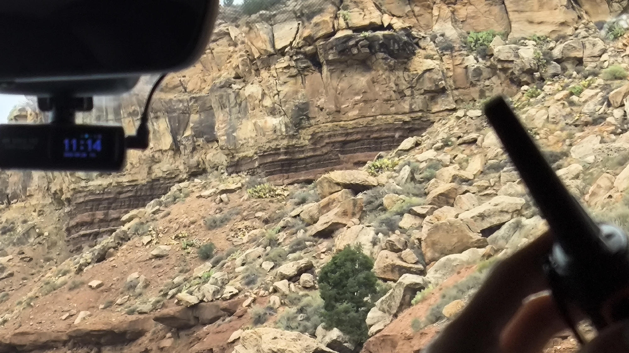 This image was taken from inside a vehicle driving through a rocky, arid landscape in Rockville, United States of America. The foreground is dominated by the interior of the car, with a dashcam visible on the rearview mirror displaying the time "11:14". A hand holding a dark object, possibly a walkie-talkie, is partially visible in the lower right corner. The background features a steep, layered rock cliff face, characteristic of desert canyons. Sparse green vegetation, including a small tree, is scattered across the rocky terrain. The overall impression is a scenic drive through a natural, rugged environment.