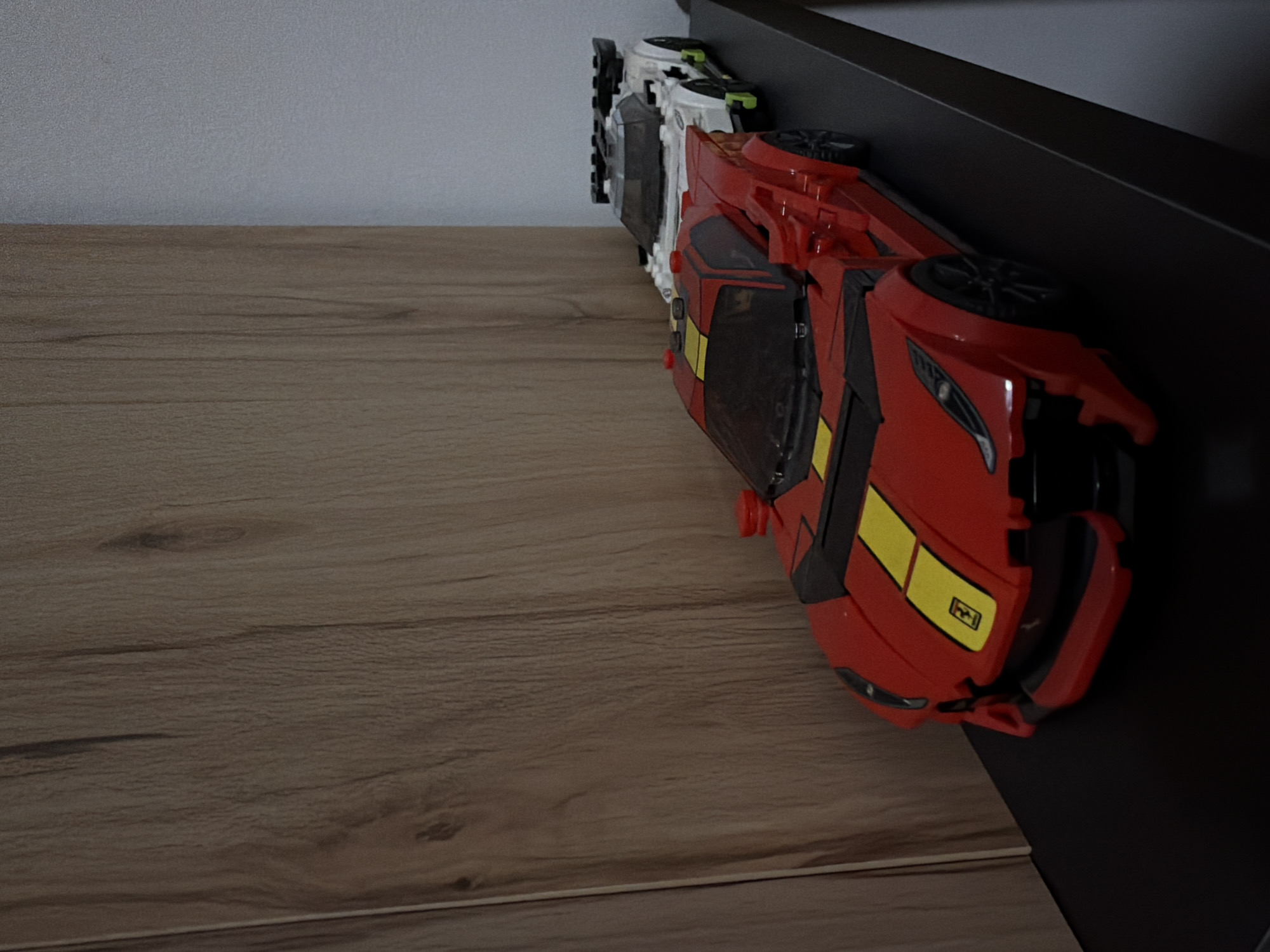 Two toy race cars are displayed on a black shelf. The car in the foreground is red with yellow racing stripes and a small yellow emblem on the hood. The second car, behind and to the right of the first, is white with green accents. The cars are positioned in front of a light brown wooden textured surface that appears to be a wall or furniture. The overall lighting suggests an indoor setting. There is no discernible information in the image that would indicate a specific location in Sankt Augustin, Germany.
