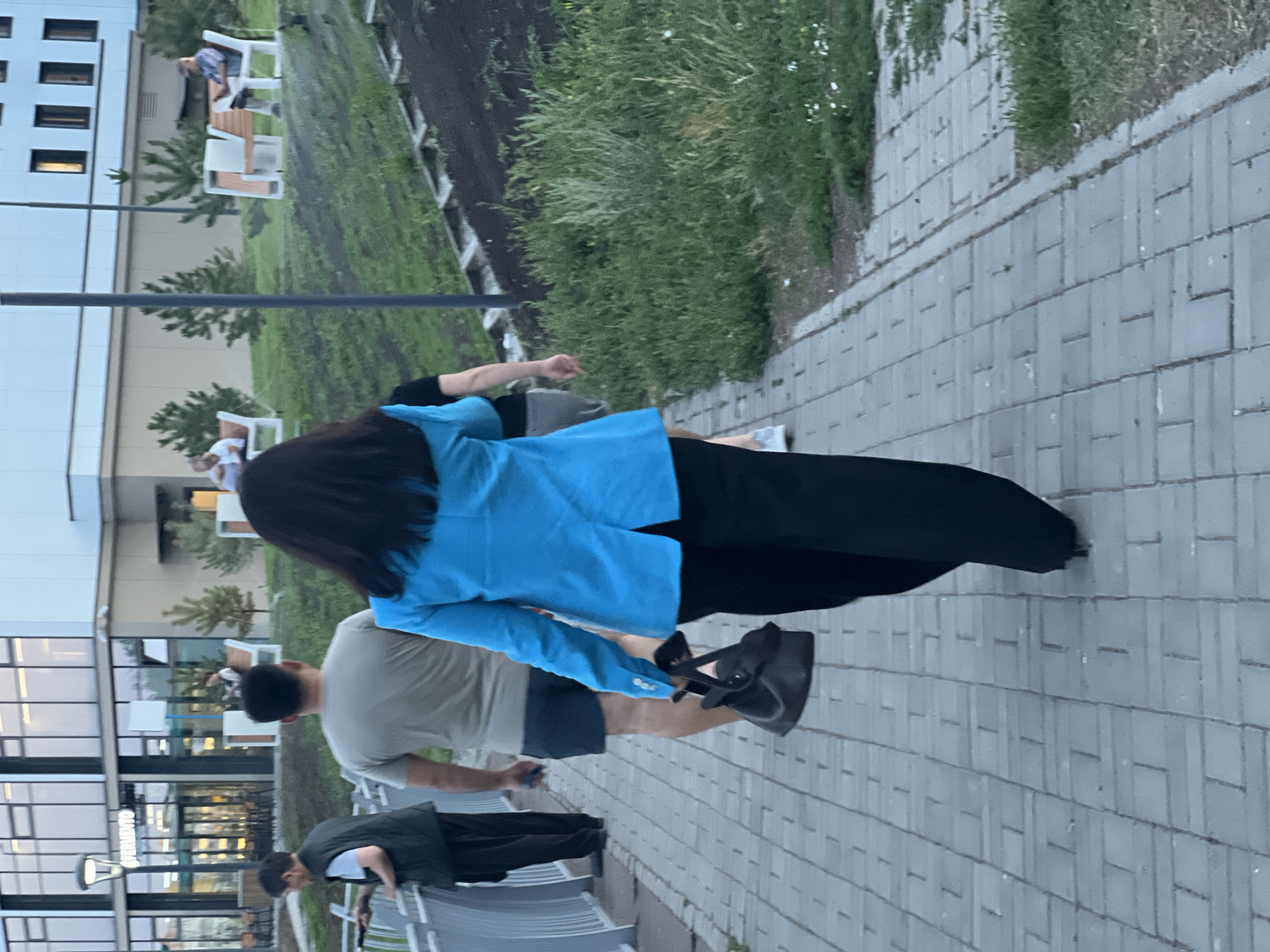 The image depicts a scene on a paved walkway in Astana, Kazakhstan. In the foreground, a person wearing a bright blue blazer and black pants walks away from the camera, carrying a dark handbag. To their left, a man in a light-colored t-shirt and dark shorts walks in the same direction. Further left, another man in a dark vest over a light t-shirt and dark pants is visible. In the background, several individuals are seated on benches in a grassy area adjacent to a modern building with large windows. The building appears to be a commercial establishment, with the word "FERMENTO" visible on its facade. The environment suggests an urban setting with landscaped areas.