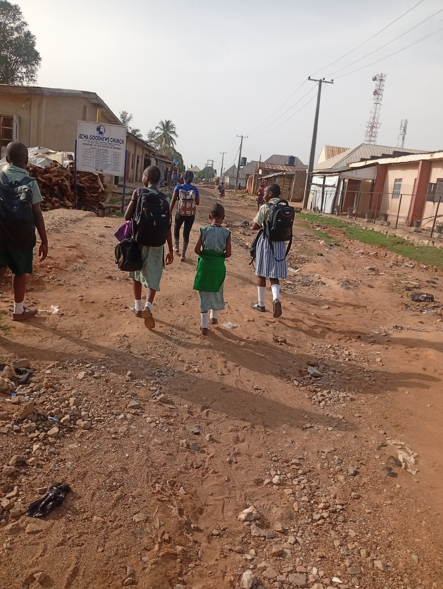 The image shows a dusty street scene in Sabon Tasha, Nigeria. Several people, primarily children in school uniforms, are walking away from the camera. They are carrying backpacks and walking on an unpaved, rocky road. On the left is a sign for "ECWA GOODNEWS CHURCH" in Kadu. Buildings line both sides of the street, and utility poles are visible overhead, along with a cell phone tower. The overall scene appears impoverished, with visible trash and debris on the ground.