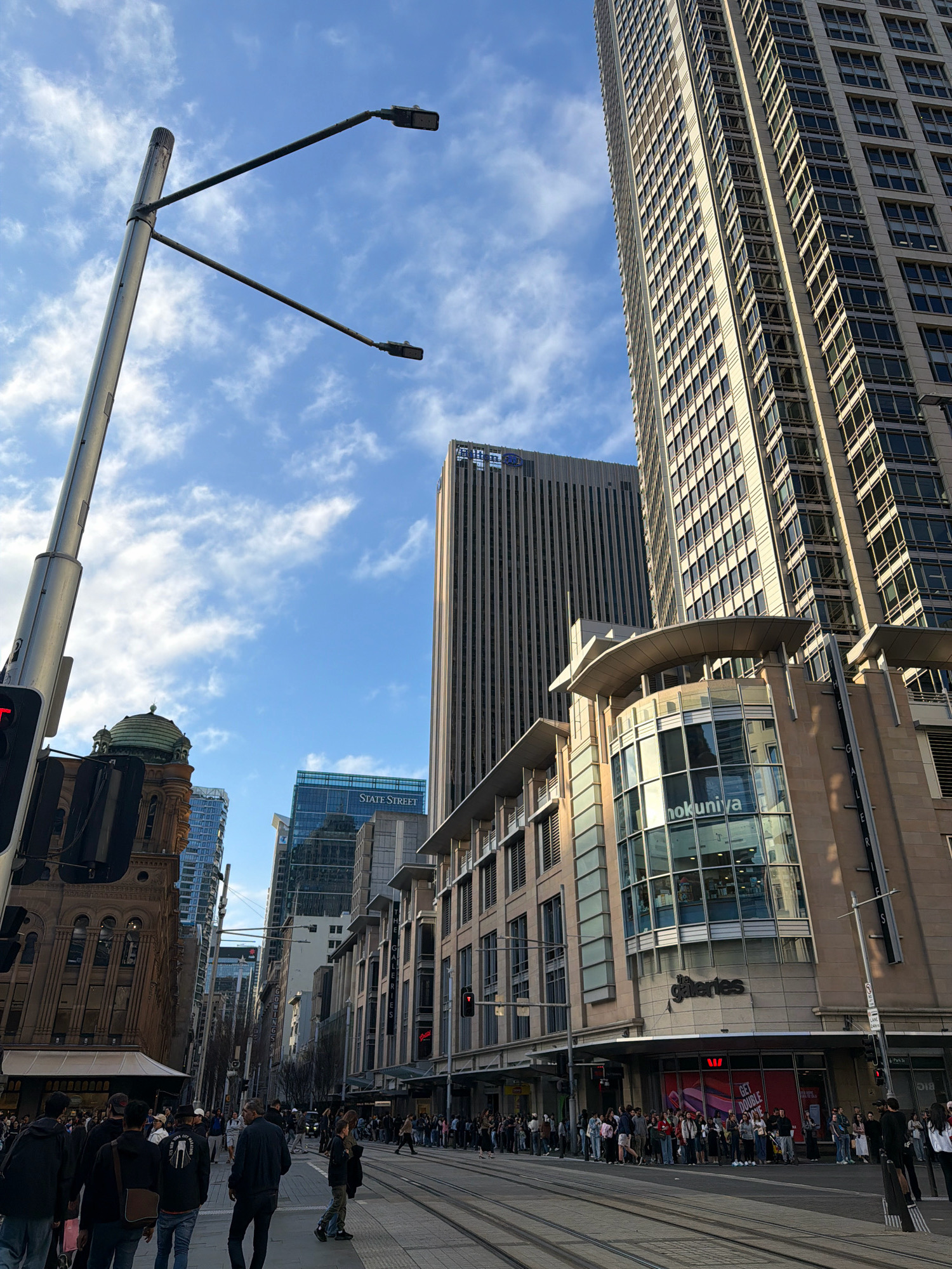 The image depicts a street scene in the Central Business District (CBD) of Sydney, Australia. The viewpoint is from street level, looking up at a variety of buildings and the sky. In the foreground, a tall, silver street light pole with two lamps extends into the frame. To the left, a portion of a building with a green dome and ornate stonework is visible.
Dominating the background are several tall office buildings. The most prominent is a light-colored skyscraper with a grid of windows. To its left is a darker, more slender skyscraper with vertical lines. Further in the distance, a blue-glass building bears the signage "STATE STREET". A modern building with curved architectural elements and large glass windows, displaying the signs "hokuniya" and "the galeries", is also a significant feature.
The street itself is paved with tram tracks. A crowd of pedestrians is gathered on the sidewalks and crossing the street, suggesting a busy urban environment. People are dressed in casual attire, with many walking or standing in groups. Some are looking towards their phones. Traffic lights are visible, indicating an active intersection. The sky is blue with scattered white clouds. The overall impression is a bustling commercial district on a bright, possibly overcast day.
