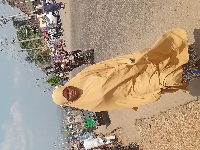 A woman in a tan hijab stands on a road. A motorcycle carrying multiple passengers moves past. Cars, trucks, and people line the street beyond. A building with a rounded roof and nearby palm tree is also visible. Utility poles run along the side of the road. The sky is pale blue. The image is rotated sideways.
