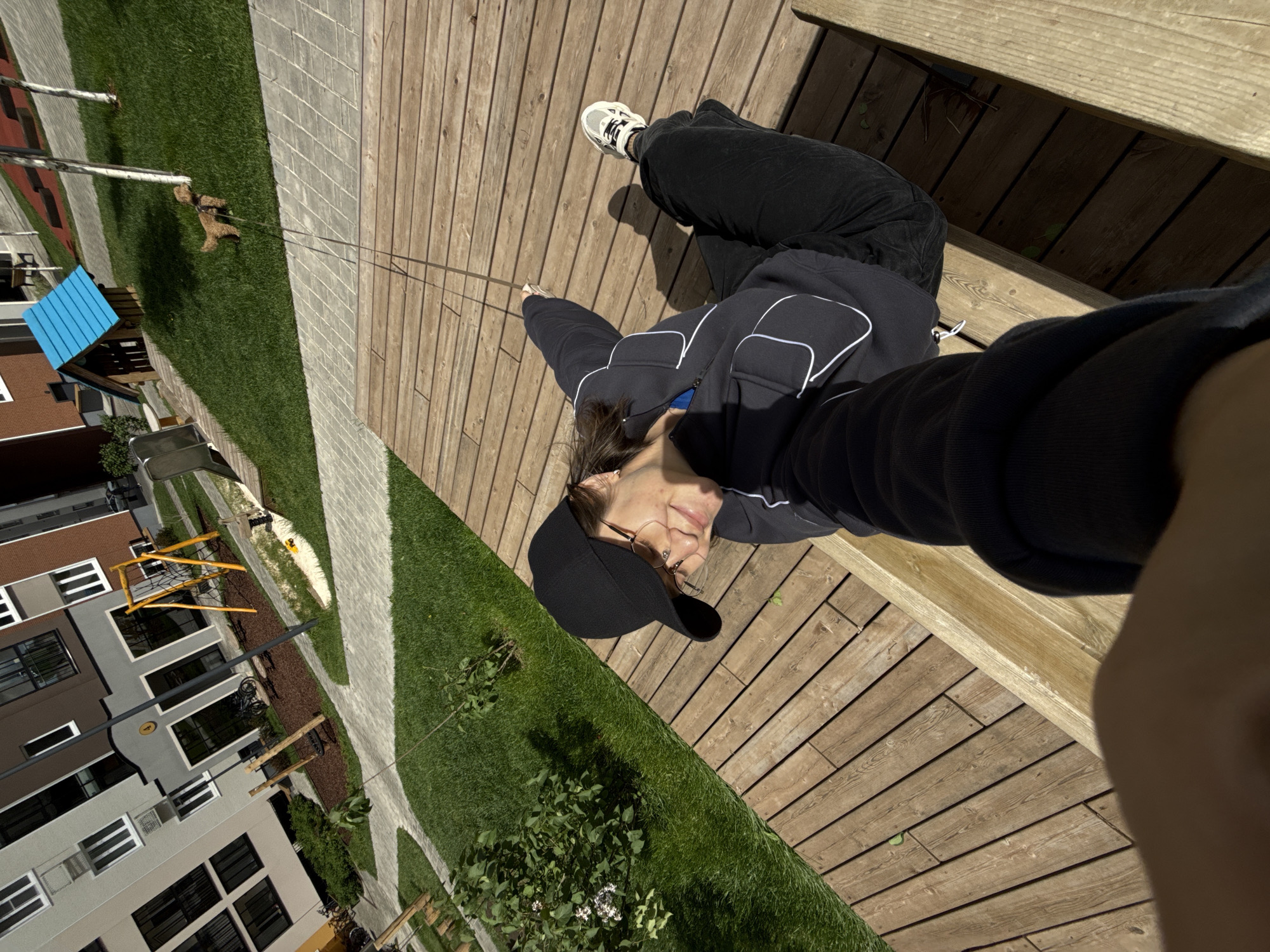 A first-person perspective captures a woman seated on a wooden structure, possibly a bench or deck. She wears a black hoodie, black pants, white sneakers, glasses, and a black baseball cap. A dog, leashed, stands on the grass in the background. The setting appears to be a public outdoor space in Izhevsk, Russia, judging from the architectural style of the buildings in the background, which are modern apartment blocks with multiple windows and brown and grey facades. A children's playground with a blue roof and climbing structures is also visible in the background. The weather appears sunny.