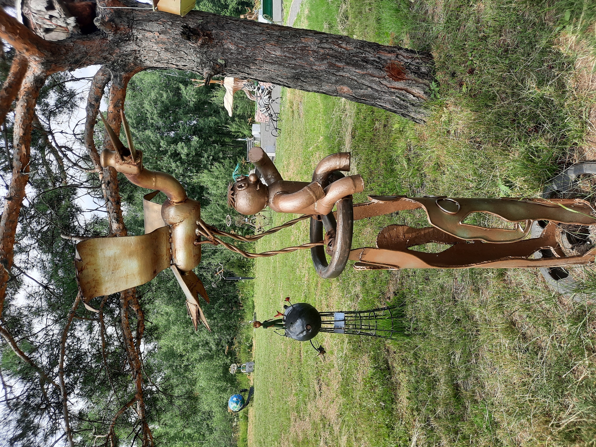In Зеленогорский, Russia, an outdoor sculpture garden features several whimsical metal artworks. In the foreground, a large metal sculpture depicts a bird with outstretched wings perched above a circular frame supported by abstract human-like figures. To the left, another sculpture shows a small figure resembling "The Little Prince" standing on a black sphere, which is itself mounted on a wire stand. In the background, a blue globe sculpture is visible, along with other indistinct metal art pieces and trees. The sculptures are situated in a grassy area with a large pine tree dominating the right side of the frame.