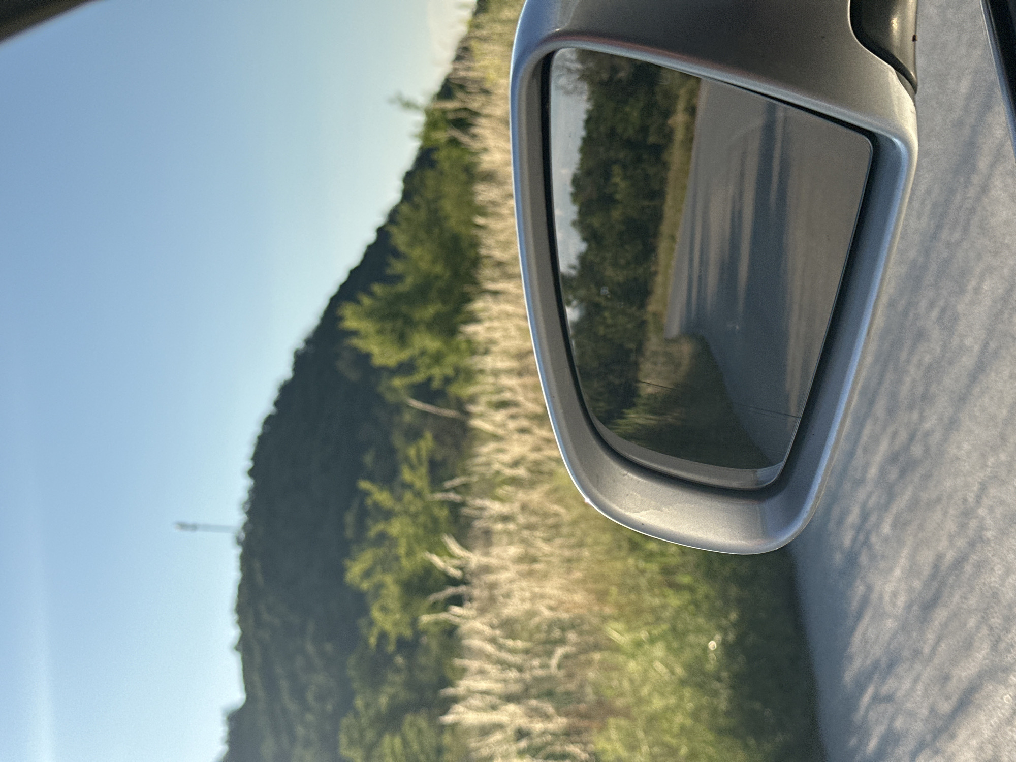 The image captures a view from a moving vehicle, focusing on a side-view mirror reflecting a winding road through a wooded area. In the background, a grassy hillside is visible under a clear blue sky. A tall communication tower stands on the crest of the hill. The foreground shows the car's exterior and the road surface, with long shadows suggesting it is late afternoon or early morning. The scene appears to be located on the outskirts of Prisovo, Bulgaria, given the landscape and vegetation.