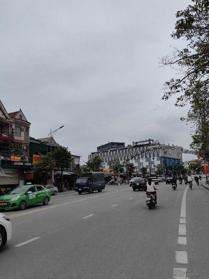 The image captures a daytime urban street scene in Trường Vinh Ward, Vietnam, under an overcast sky. The foreground shows a multi-lane asphalt road, marked with a solid double yellow line for center separation and dashed white lines delineating lanes.
On the left side of the road, a green Hyundai i10 taxi is present, followed by the front bumper of a white car. Ahead, a blue cargo truck with a canvas cover on its bed is in motion. Buildings on the left are multi-story structures with varied architectural styles, including one prominent pink-toned building with a gabled roof and a balcony adorned with flowering plants. Vietnamese signage, such as "CHỢ PHÚC VINH" and "TRƯỜNG," is visible on these commercial/residential facades. Overhead power lines and a streetlight are also present.
On the right side of the road, multiple motorcycles and scooters are in various positions, some ridden by individuals wearing helmets. A black sedan and another dark-colored vehicle are also observable. The background is dominated by a large, modern building with a distinctive facade composed of repeating gray and white square/cross patterns and blue-tinted glass sections. Signage on this building includes "ANH" and "CGV." An elevated structure, potentially a pedestrian bridge or decorative archway, spans across the street from this building, featuring additional red banners and flags. Trees with green foliage line both sides of the street, and red banners/flags are affixed to poles along the right sidewalk.