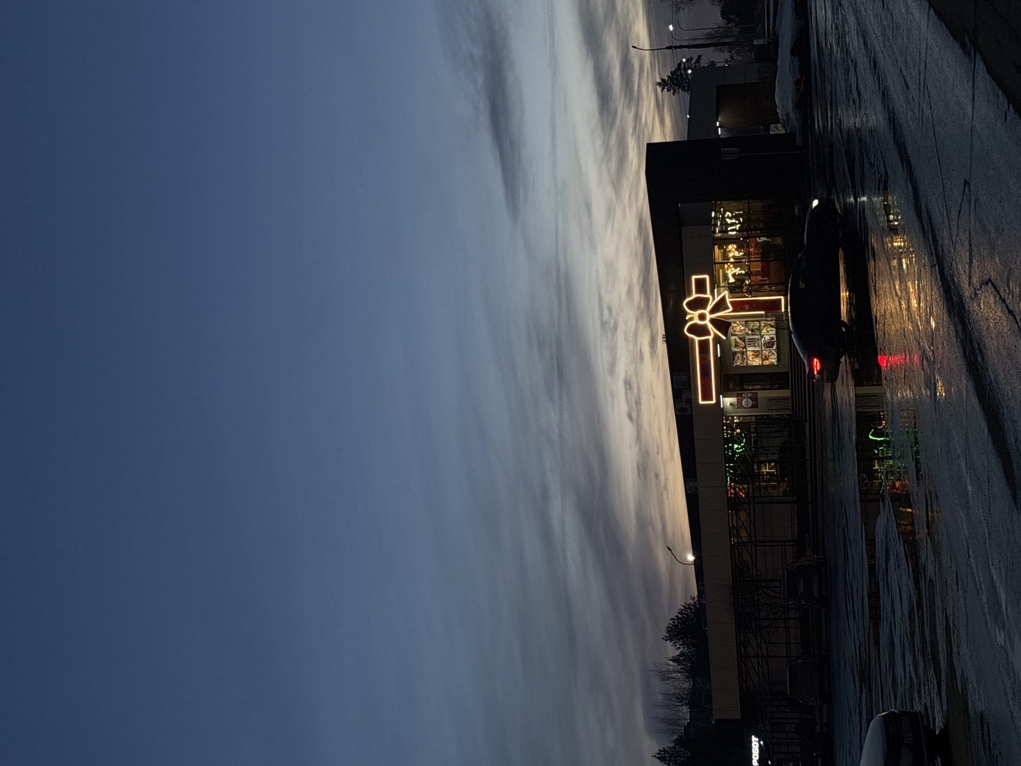The image depicts an outdoor scene in Chaikovsky, Russia, during dusk or early night. A single-story commercial building with a dark-colored facade and extensive glass windows is centrally positioned. On its front, a large, bright orange-yellow outline of a gift bow is illuminated. Through the windows, internal lighting reveals various retail displays, including partial text such as "JAM" and "РОБОТ" (ROBOT).
The foreground features a dark, wet asphalt parking lot, with numerous puddles reflecting the building's lights and the red taillights of a dark-colored car in the mid-ground, suggesting it is either stopped or moving slowly. Other dark vehicles are partially visible parked further back and on the far left. Patches of snow are present on the ground to the right of the building. In the background, a line of bare deciduous trees and dark evergreen trees are silhouetted against the sky. The sky transitions from a dark blue overhead to a lighter grey-orange hue near the horizon, exhibiting elongated, horizontal cloud formations. Streetlights are visible, contributing to the artificial illumination. No individuals are discernible in the frame.