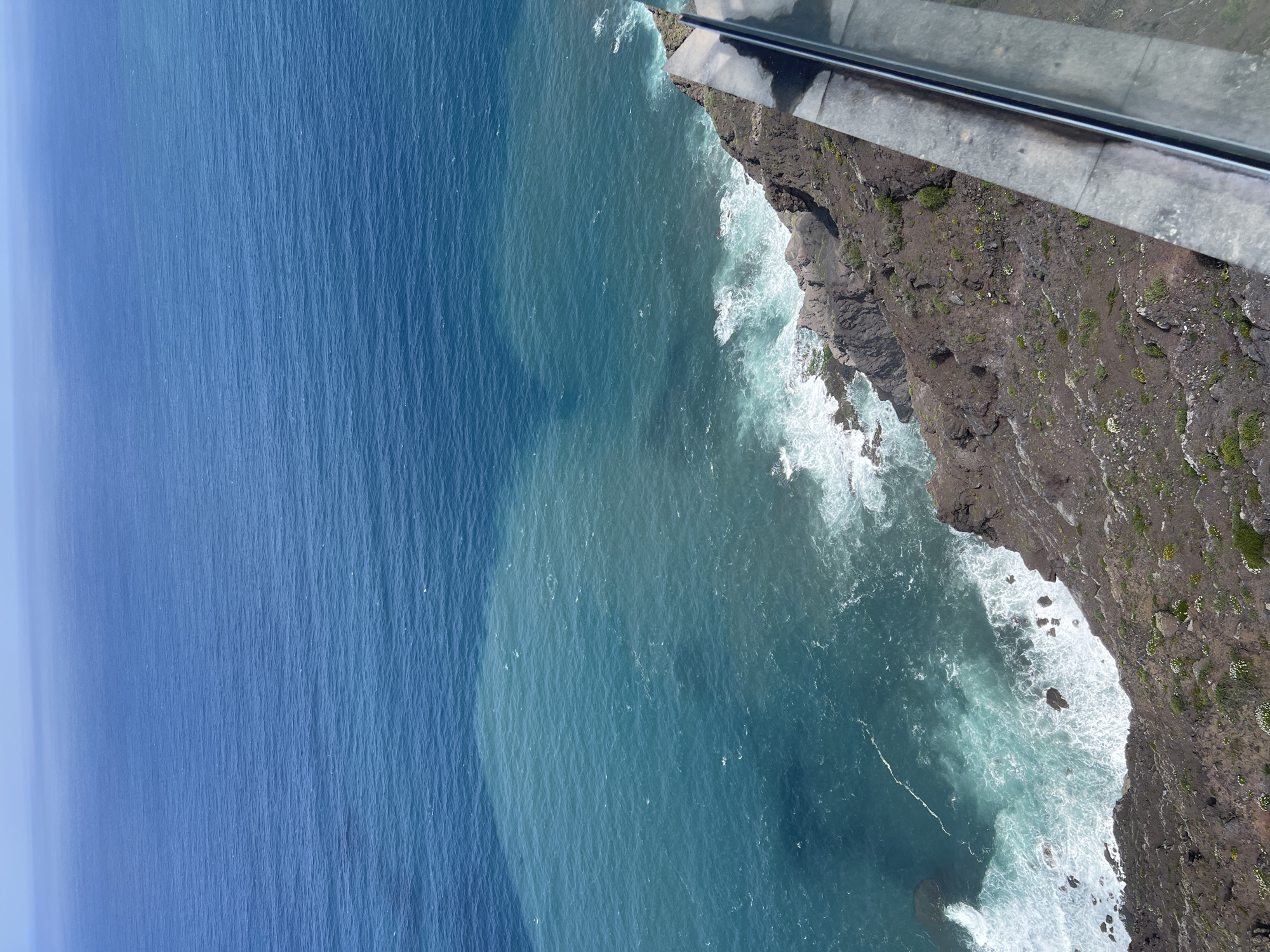 The image captures an aerial view of the coastline in Tirma, Spain. To the left, the deep blue of the ocean stretches out, contrasting with the lighter turquoise water closer to the shore. The rocky coastline, characterized by dark volcanic rock, is visible on the right, with white waves crashing against it. Vegetation is sparsely scattered along the cliffside. A portion of a glass platform is visible in the upper right corner.