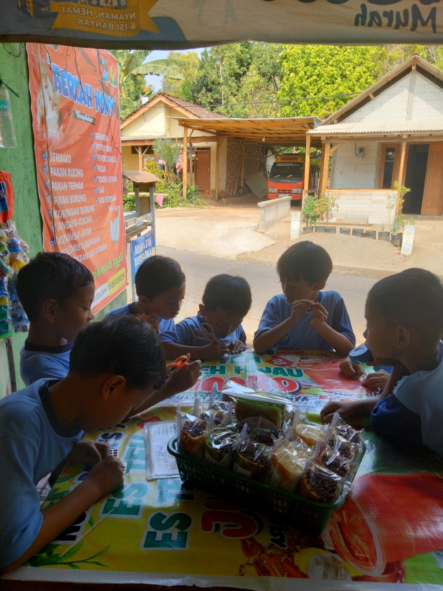 A group of five young boys are gathered around a table outdoors, in what appears to be a storefront in Sidorejo, Indonesia. The table is covered with a colorful, patterned tablecloth featuring cartoonish images and text, some of which is in Indonesian. In the foreground, a green plastic basket contains several individually wrapped snacks, seemingly sweet treats like cakes or pastries, with visible chocolate sprinkles. Some of the boys are looking down at the table, possibly interacting with the snacks or engaged in an activity. One boy in the center is holding a small, dark object between his fingers, while another to his right is holding orange-handled scissors. To the left, a boy with dark hair looks intently at the table. In the background, there is a building with a white brick facade and a wooden porch, and a red truck is visible under a covered outdoor area. A large orange banner with Indonesian text and images of animals is hanging on the left side of the frame, suggesting this is a shop or stall. The environment appears to be a rural or semi-urban area, with trees and vegetation visible behind the buildings. The lighting indicates it is daytime.