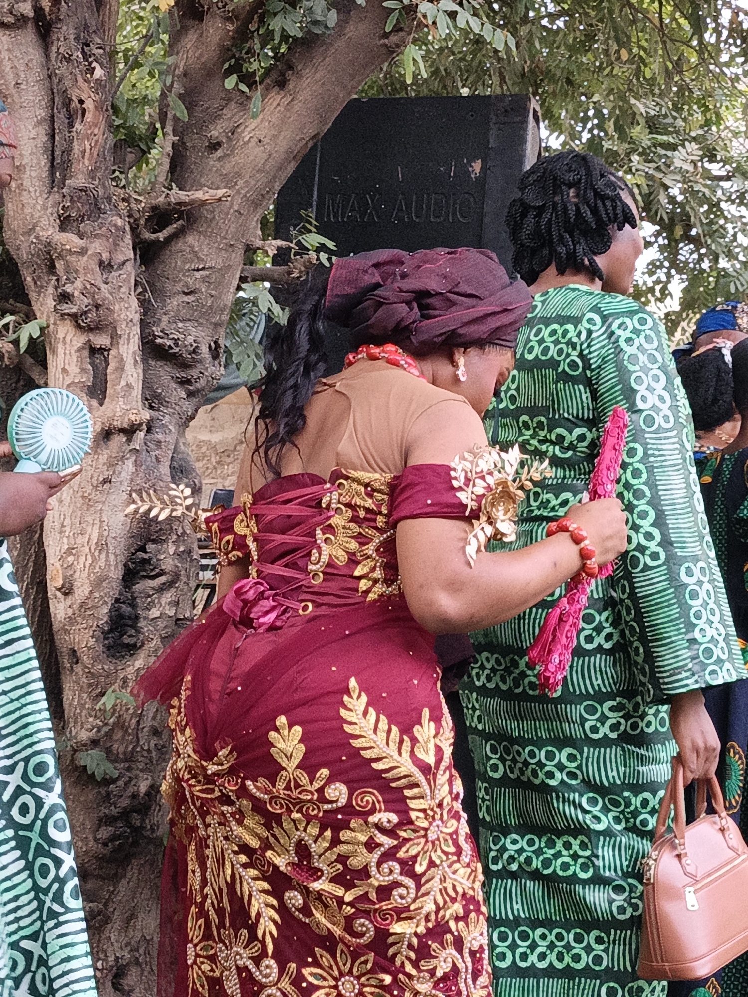 This medium shot captures two women and parts of others at an outdoor social gathering in Gafan, Nigeria, during daylight hours. The woman in the foreground wears a striking dark red traditional dress adorned with elaborate gold embroidery and lacing on the back. She pairs this with a matching headwrap, red beaded jewelry (necklace and bracelet), and holds a decorative pink fan. Beside her, another woman is partially visible from the back, dressed in a vibrant green traditional patterned gown, carrying a brown handbag, and sporting dark braided hair. A third person on the far left holds a light blue portable fan, suggesting warm weather. In the background, a large tree trunk dominates the left side, while a black speaker box with the text "MAX AUDIO" is visible on the right, indicating music or announcements are part of the event. The festive attire and outdoor setting suggest a celebration or community gathering.