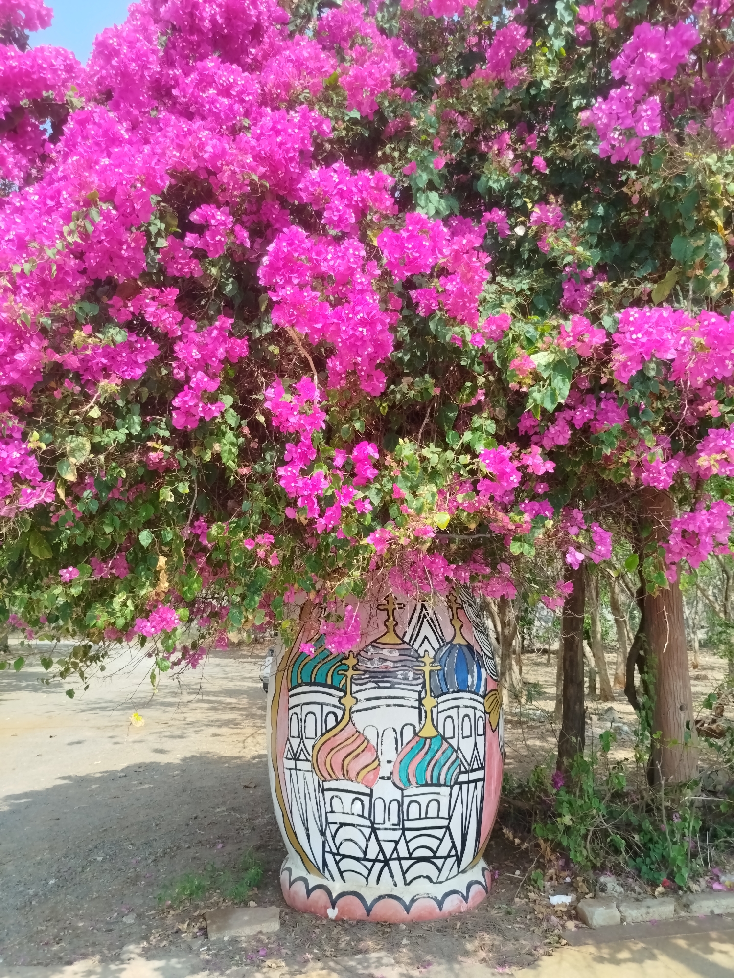 A vibrant pink bougainvillea bush overflows from a large, painted pot resembling a matryoshka doll. The pot features illustrations of Russian Orthodox churches with colorful domes. The scene is bathed in daylight, and the ground around the pot is a mix of pavement and dry earth scattered with fallen pink blossoms. A slender tree trunk stands to the right of the pot. The location is likely Ho Chi Minh City, Vietnam.