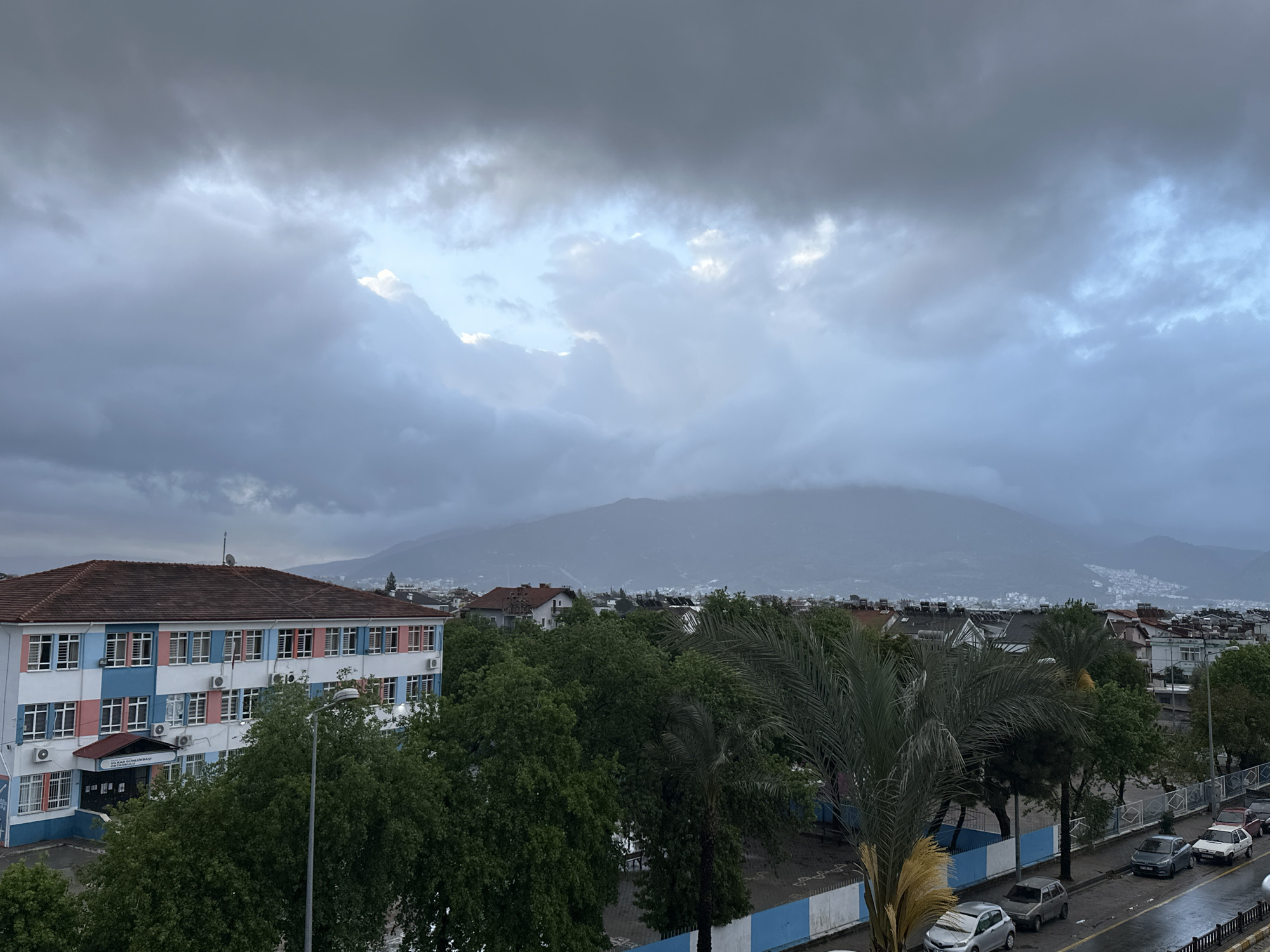 The photograph shows a cityscape in Fethiye, Turkey under a cloudy sky, likely during or after rainfall, as the road appears wet. A large building with a brown tile roof, painted in blue, white, and pink, stands on the left side of the frame; its sign reads "ILKAR ÖĞRETMOKASI" (likely a misspelled version of "İlkokul Öğretmenevi"). Several cars are parked along the street on the right side of the frame. The trees are dense and green, with a palm tree visible in the foreground. The background features a mountain partially obscured by clouds.