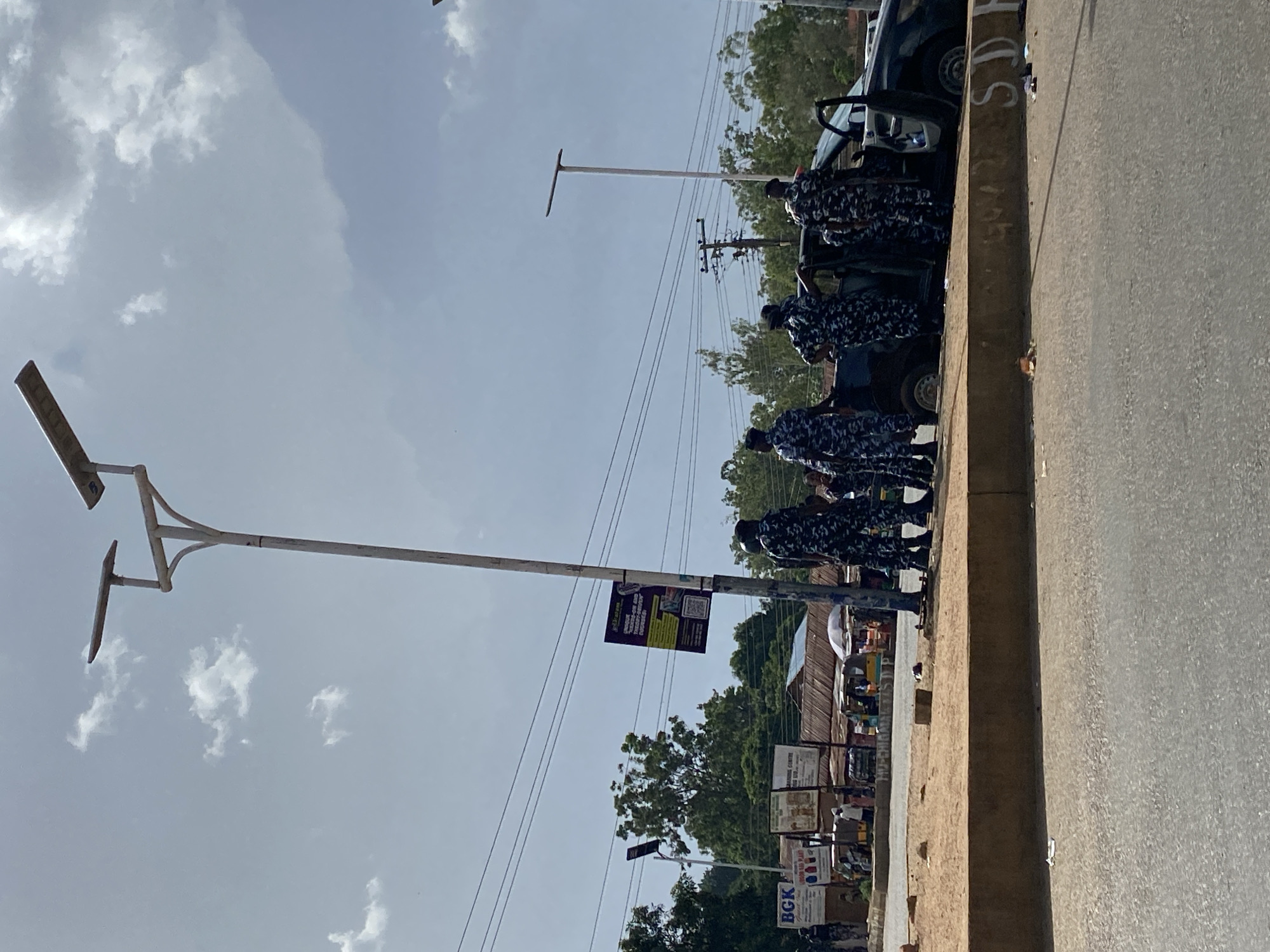 In Bukuru, Nigeria, a group of individuals wearing camouflage uniforms are gathered around an open-doored dark-colored vehicle. They appear to be law enforcement officers. One of the officers stands near a lamp post that has a poster attached to it, displaying text and a QR code. The scene is outdoors, with a road in the foreground and a sidewalk or embankment separating it from a background that includes shops, trees, and utility poles with power lines. The sky is partly cloudy.