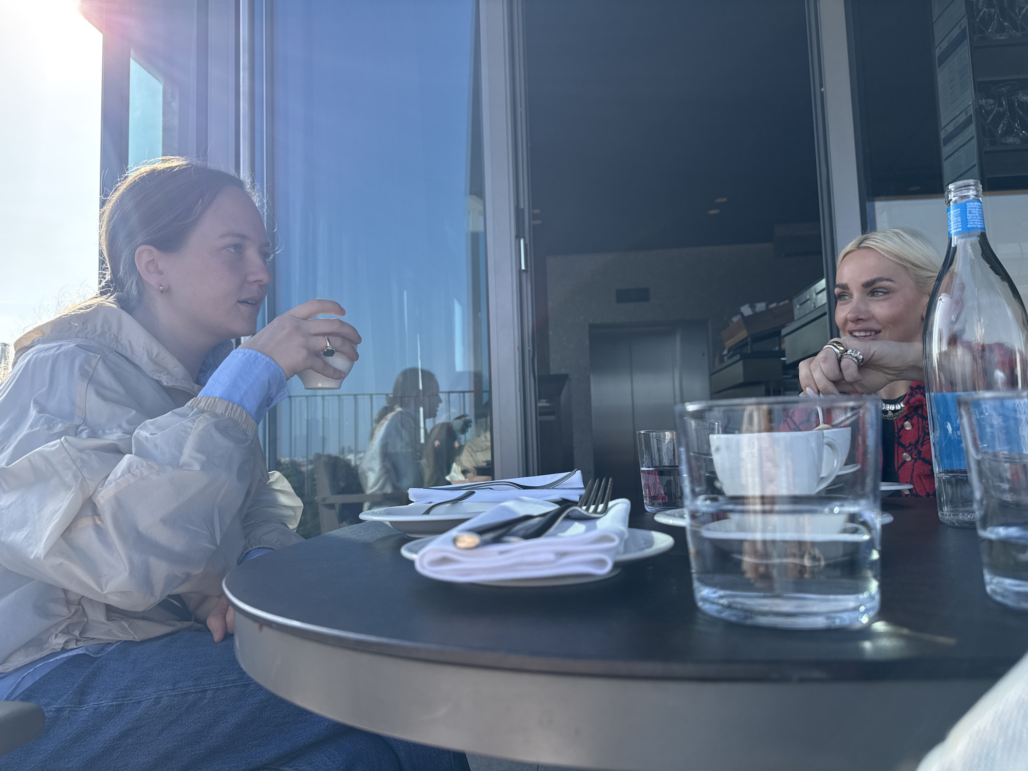 Two women are seated at a round table in an indoor setting, presumably a restaurant in Milan, Italy. The woman on the left is holding a white cup, possibly a coffee cup, while looking towards the right. She is wearing a light-colored jacket over a blue shirt and blue jeans. The woman on the right is looking towards the left and has her hand near a white cup and saucer. She has blonde hair and is wearing a red and black patterned jacket.
On the table, there are multiple water glasses, silverware wrapped in white napkins on white plates, and a clear glass bottle filled with water. In the background, there are large glass windows offering a view of the city, as well as an elevator and other interior elements of the restaurant, suggesting it's located on a higher floor or rooftop. The sunlight is strong. The overall impression is of a relaxed conversation during a meal or coffee break.