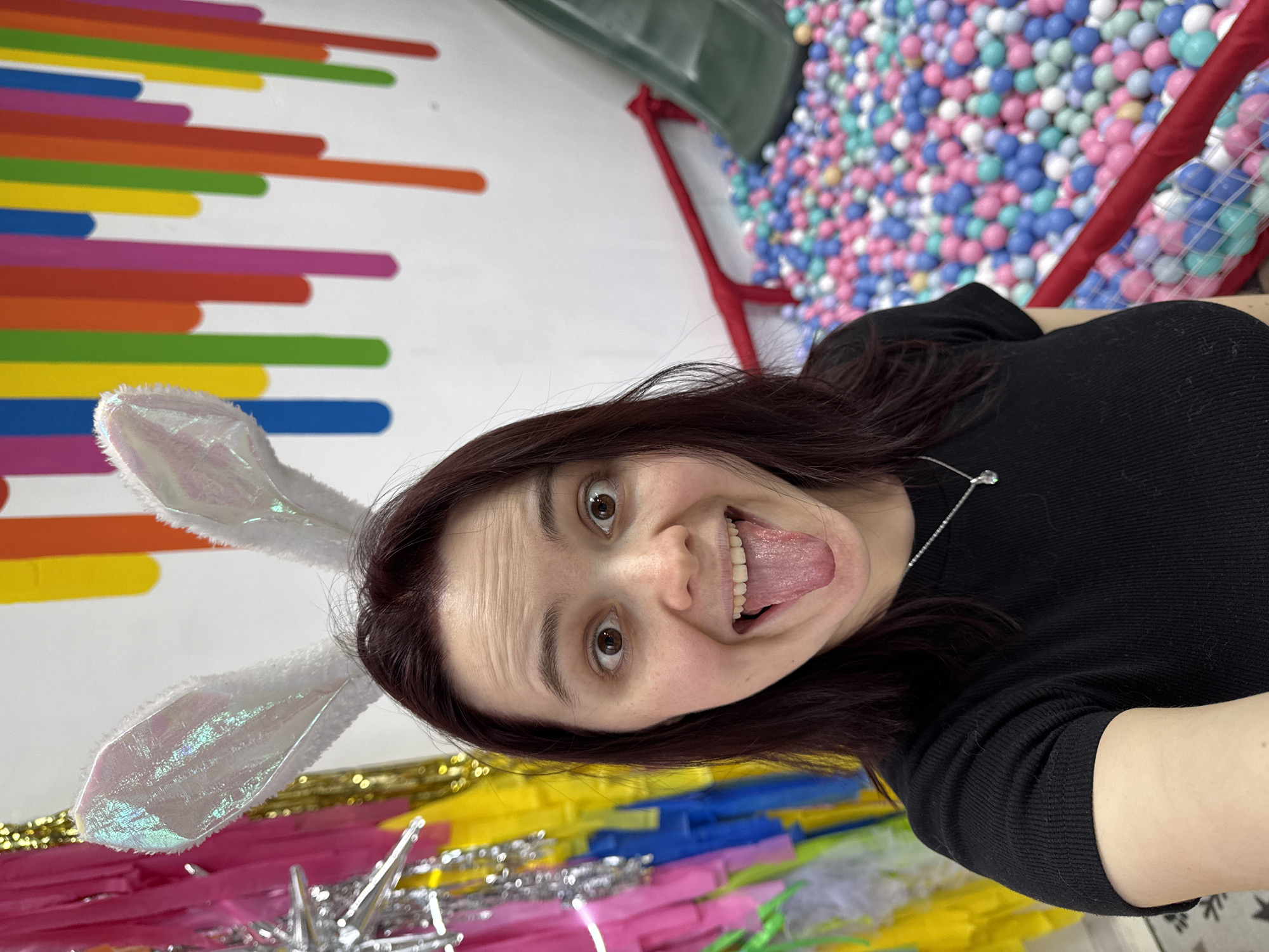 A woman wearing rabbit ears poses for a selfie. She has brown hair, fair skin, and is wearing a black shirt and a silver necklace. Her tongue is sticking out. Behind her is a multicolored wall design with horizontal bars, as well as a ball pit filled with blue, pink, and white balls. The bottom of the frame shows colorful streamers. The photograph was likely taken in a play area in Izhevsk, Russia.