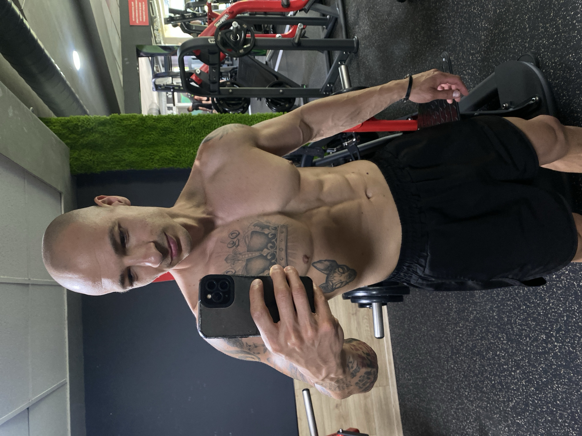 A bald, shirtless man with tattoos on his chest and arm, wearing black athletic shorts, takes a selfie in a gym. He is holding a smartphone in his right hand. In the background, various gym equipment, including weight machines and dumbbells, are visible. The gym has black walls and a textured floor. A vertical section of artificial green grass is mounted on a wall. The scene takes place in the city of Pleven, Bulgaria.