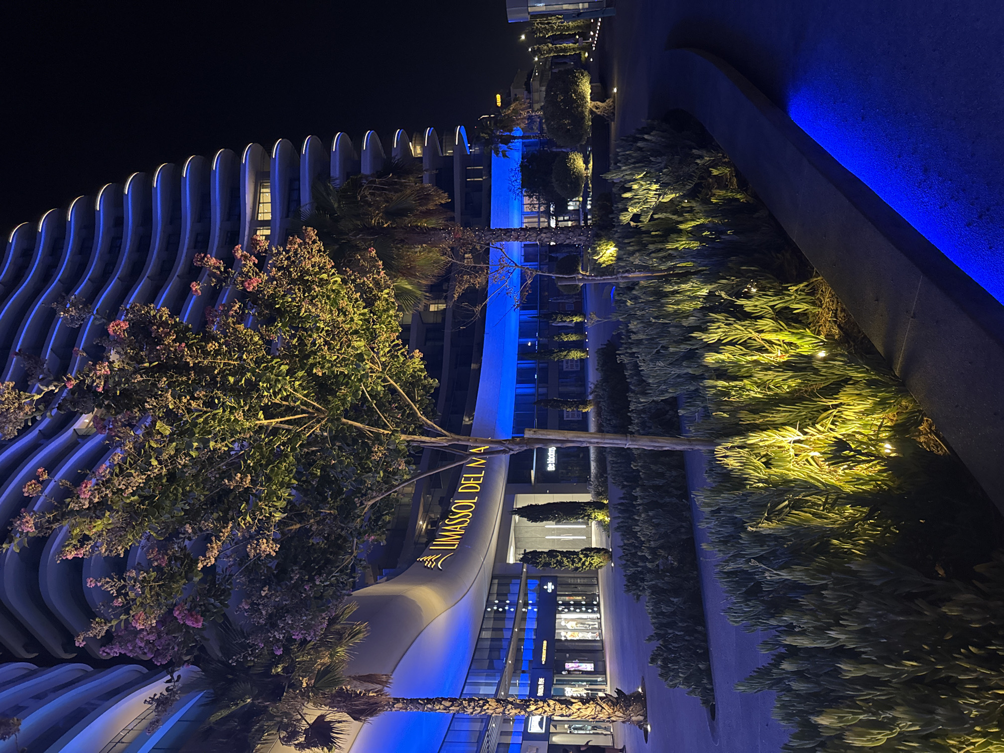 An exterior, night-time, eye-level, medium shot captures the Limassol Del Mar complex in Germasogeia Municipality, Cyprus. The building, characterized by its undulating, multi-story balconies illuminated with blue neon lights, dominates the background. In the foreground, a lush garden area features dense, green shrubbery, some of which is illuminated by warm, yellow spotlights. A slender tree with scattered pink blossoms stands in the mid-ground, partially obscuring the building. Two palm trees are visible to the left, with one taller palm to the far left and a shorter one further back. The ground in front of the garden is a dark asphalt path, bordered by a raised concrete curb that also emits a blue light. The entrance to the complex, marked with the "LIMASSOL DEL MAR" logo in yellow lettering, is visible on the left, featuring storefronts with illuminated displays and glass facades. The overall scene conveys a modern, luxurious, and architecturally striking urban environment.
