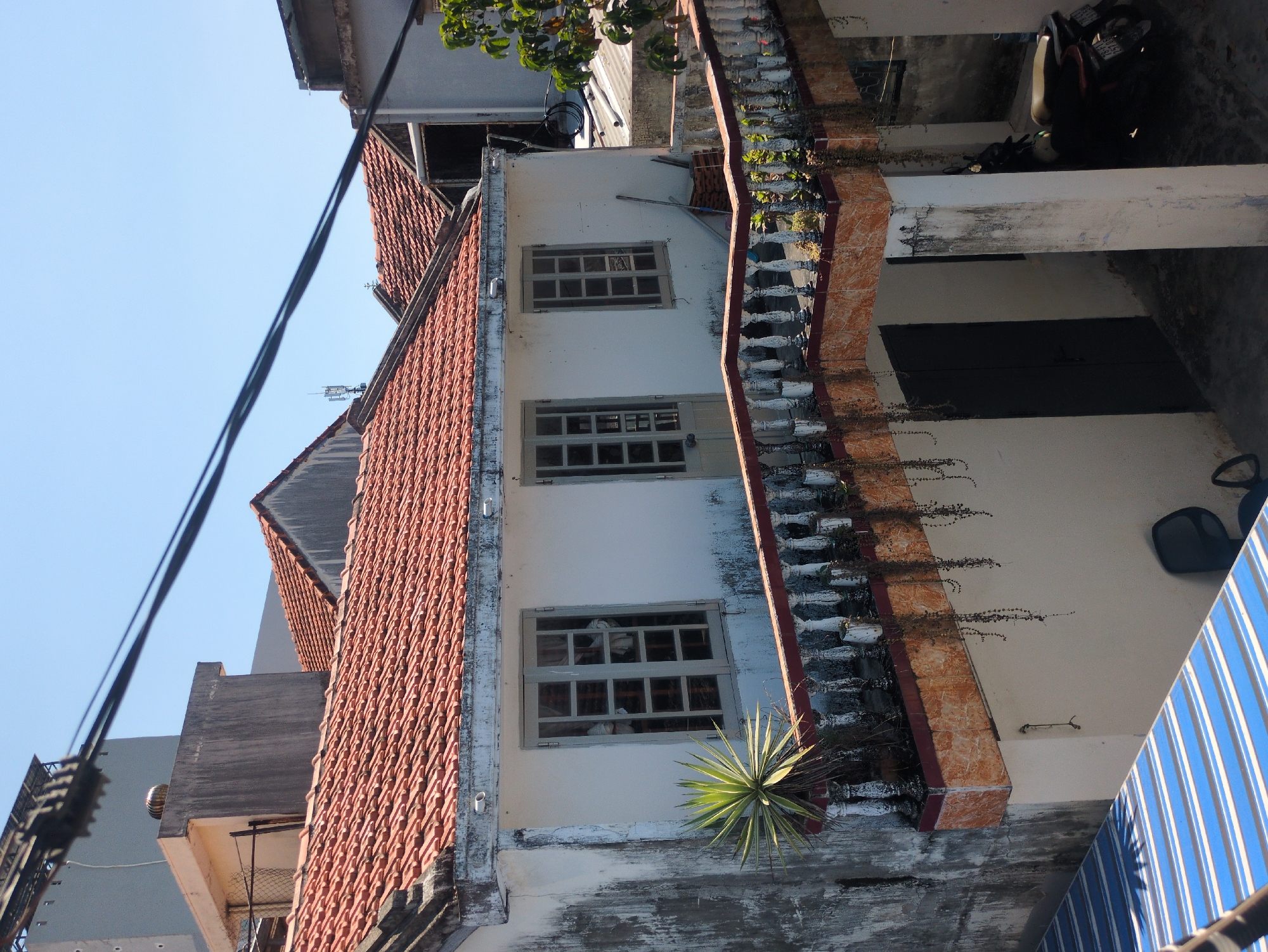 The image shows a building exterior in Tuy Hòa, Vietnam. The building has a red tile roof, a white facade, and a concrete railing with decorative white balusters and a red-brown tiled top. There are three windows on the facade, each with multiple panes. A spiky plant grows near the railing on the lower right. To the right of the building is an open area with a dark door, a white pillar, and a parked scooter in the background. A blue and white striped awning is visible at the bottom right. Power lines are seen against the blue sky in the upper left.