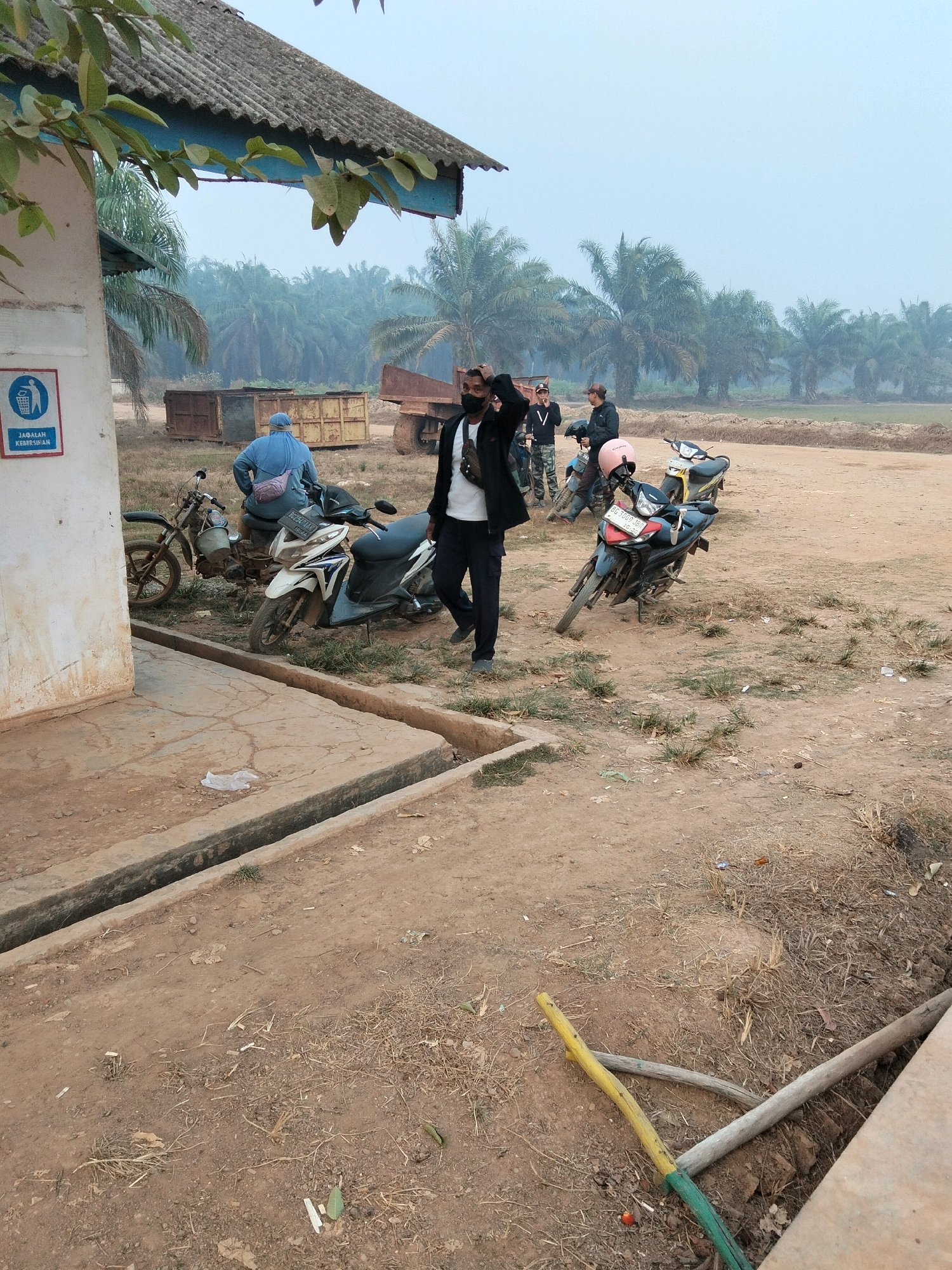 The image depicts an outdoor scene in Teluk Tenggirik, Indonesia, characterized by a dusty, rural environment with palm trees in the background, indicating a tropical or sub-tropical climate. A building with a weathered, corrugated roof and a blue overhang is partially visible on the left. A sign with a trash can icon and text in Indonesian, "JAGALAH KEBERSIHAN," meaning "MAINTAIN CLEANLINESS," is affixed to the building's exterior.
In the foreground, a concrete structure with a drainage channel runs horizontally. Several motorcycles are parked on the dirt ground. One individual, wearing a black jacket and pants, is standing near a motorcycle, with their hands near their face. Another person, wearing a blue head covering and light blue top, is seated on a motorcycle. Other individuals are visible in the background, some standing near motorcycles.
The ground is composed of dry dirt and scattered dry vegetation. In the far background, more palm trees create a dense backdrop. There are also large, rusty metal containers and a truck visible, suggesting an industrial or agricultural operational area. The overall atmosphere appears to be somewhat hazy or misty, common in certain weather conditions in tropical regions. Two wooden poles, one yellow and green and another gray, lie on the ground in the lower right corner.