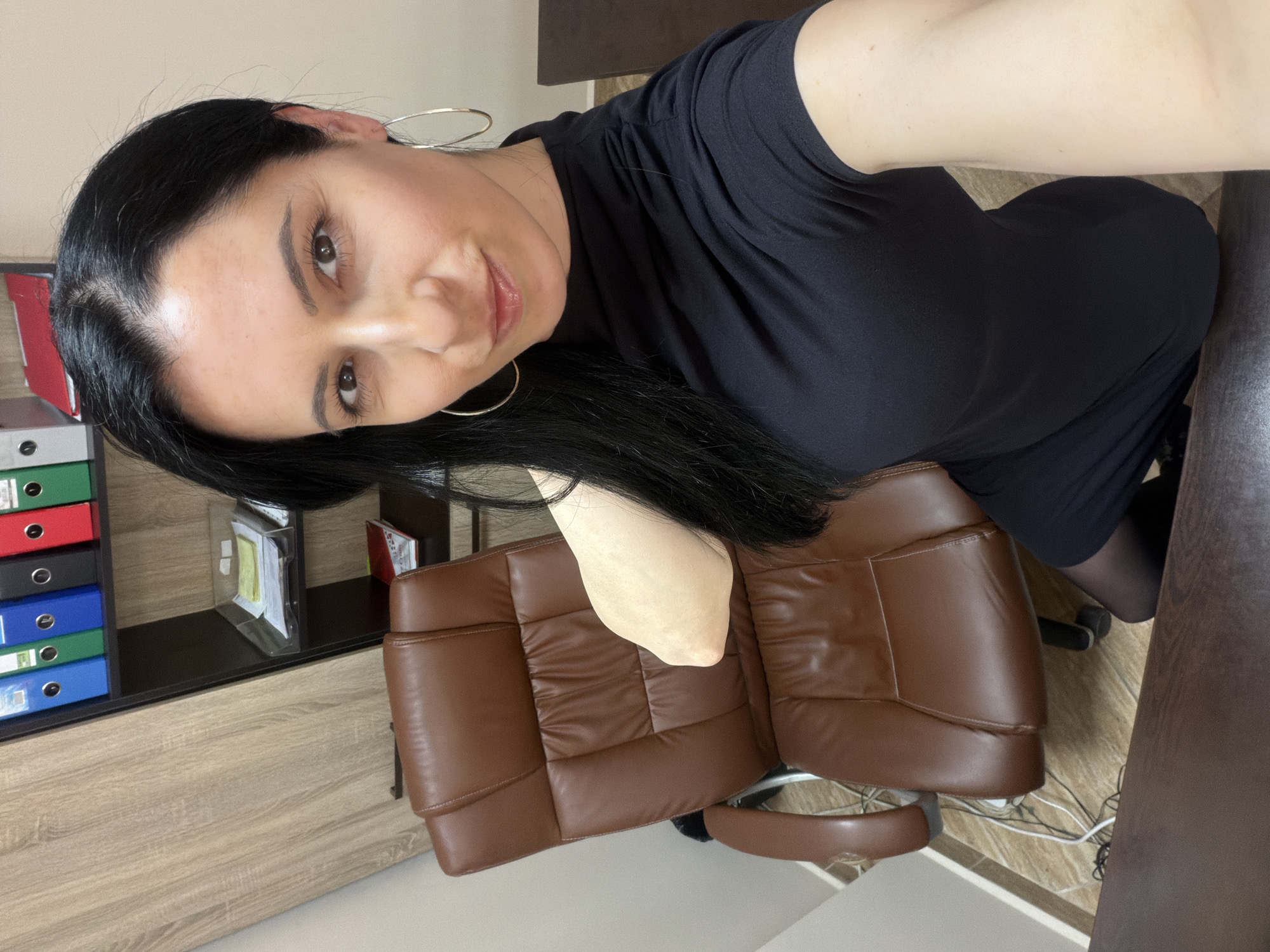A woman is in what appears to be an office. She is wearing a black dress and hoop earrings. She is sitting in a brown leather office chair. Behind her is a desk with a shelf containing several binders. It is not possible to verify the location in Pleven, Bulgaria, based on the visual information.