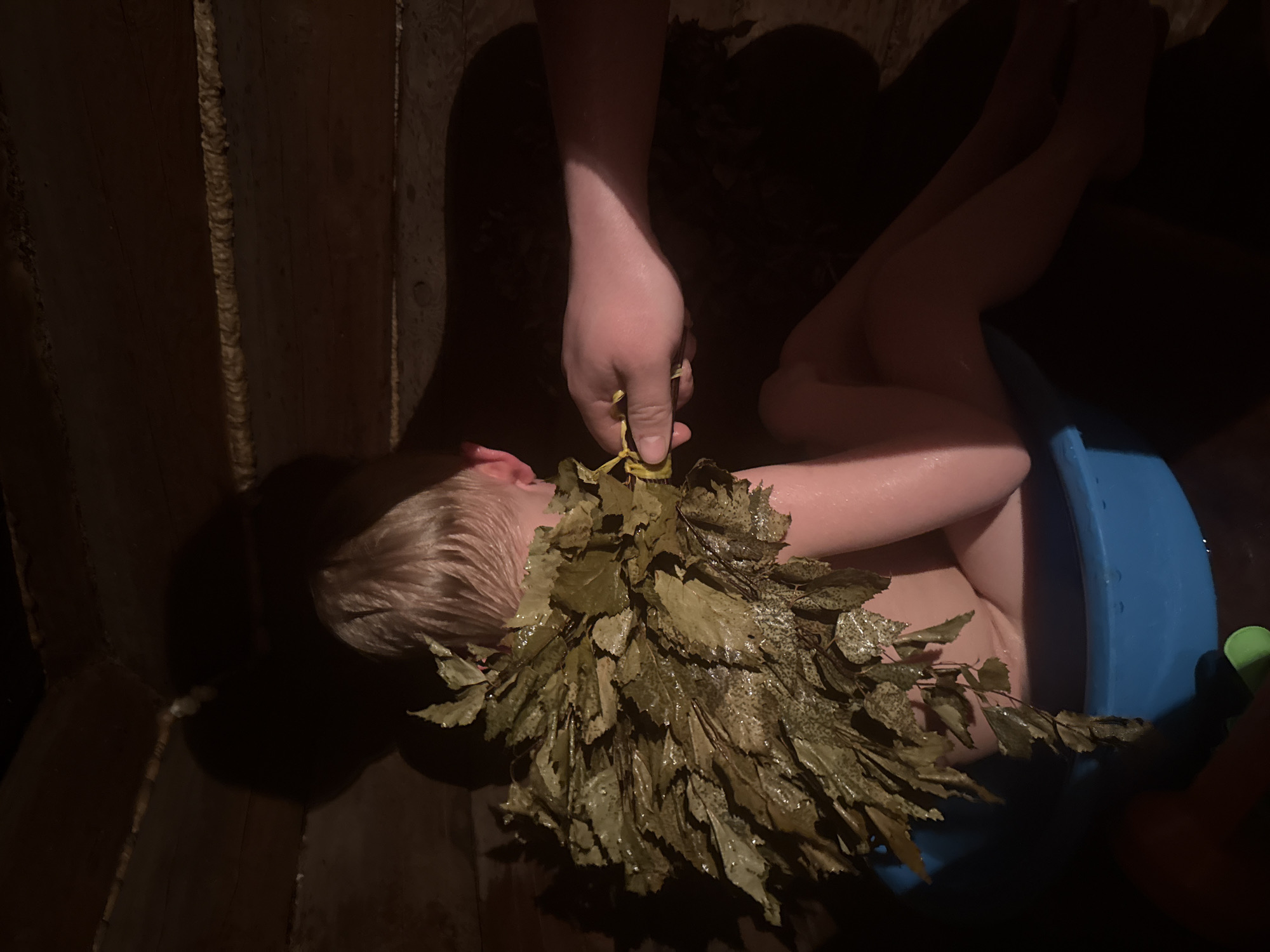 The image shows a person in a wooden structure, possibly a sauna or bathhouse, located in Luzhkovo, Russia. A child with light hair is seated in a blue tub. An adult hand holds a bundle of leafy branches, appearing to scrub or cleanse the child. The lighting is dim.